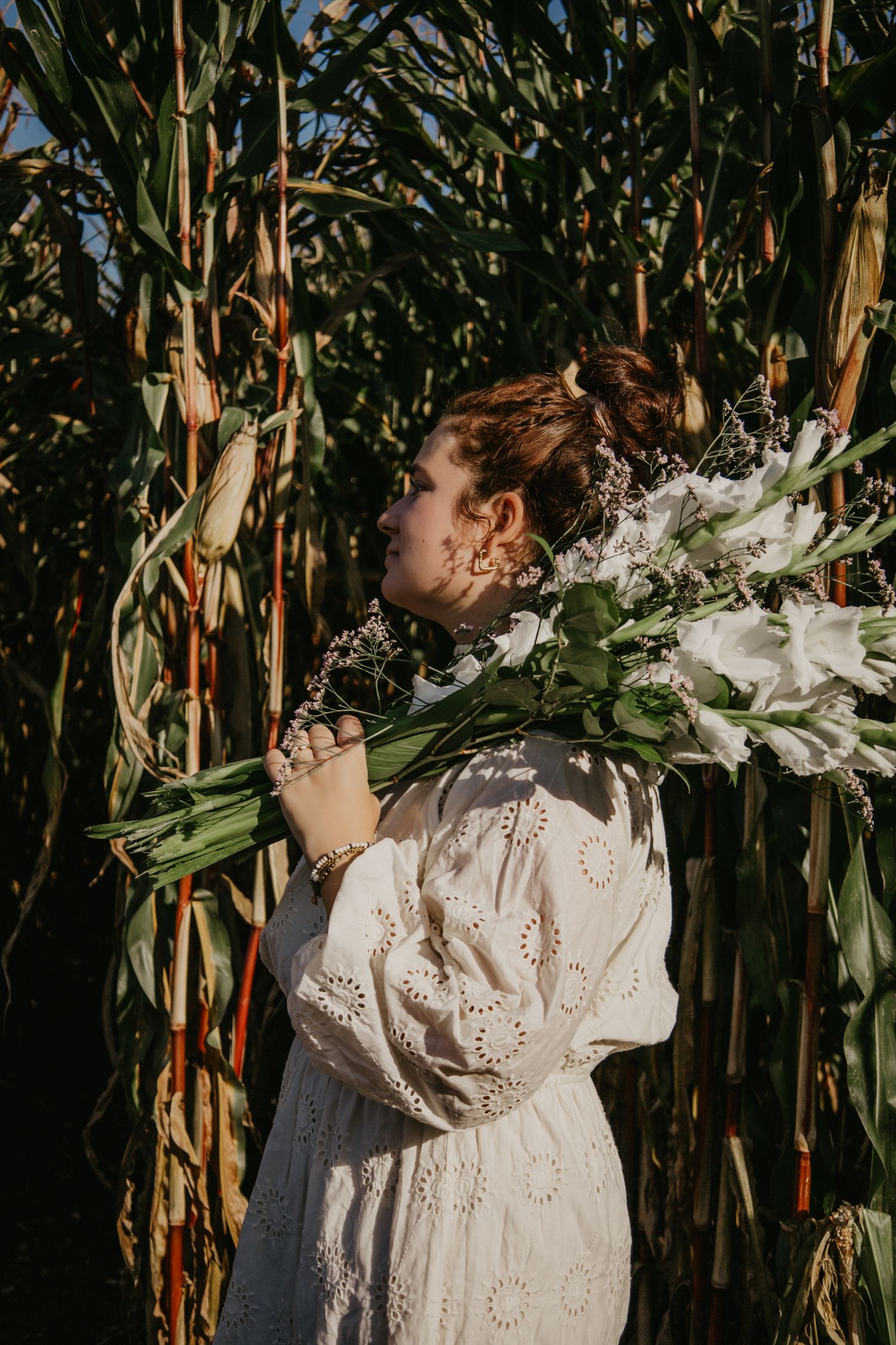 Figure in white eyelet dress holds a bouquet against a backdrop of corn stalks.