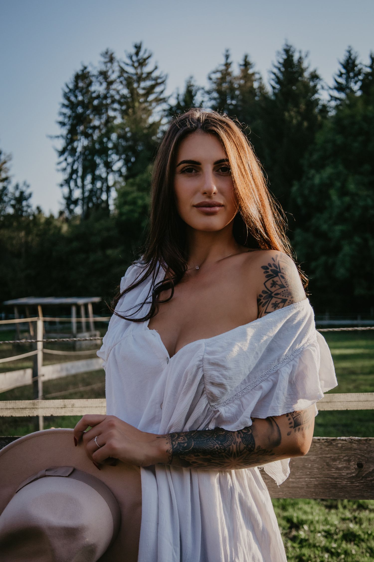 Romantic portrait in white dress showing tattoo details against rustic farm backdrop.