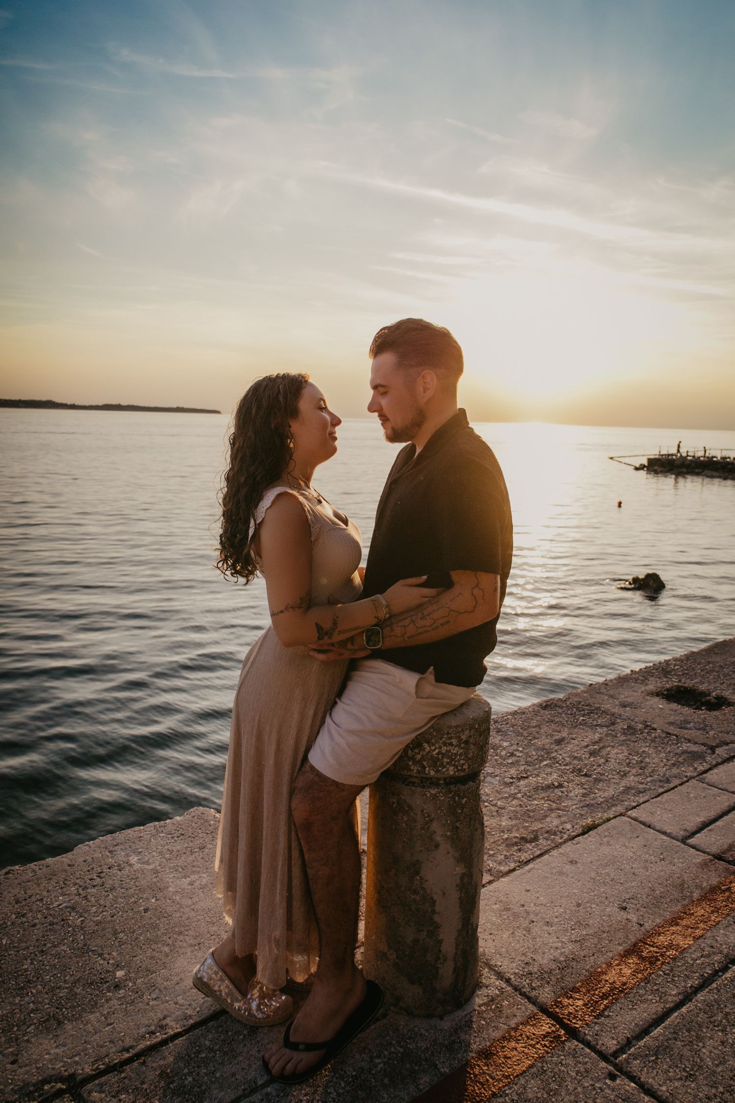 A couple embraces on a stone pier during sunset with the ocean stretching to the horizon.
