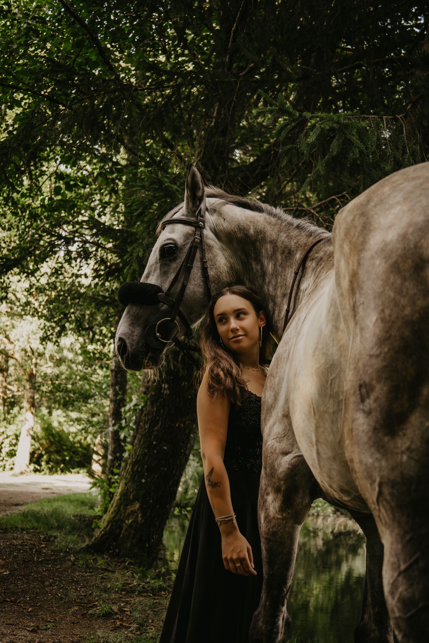 Someone in a black dress stands beside a grey horse on a tree-lined path.