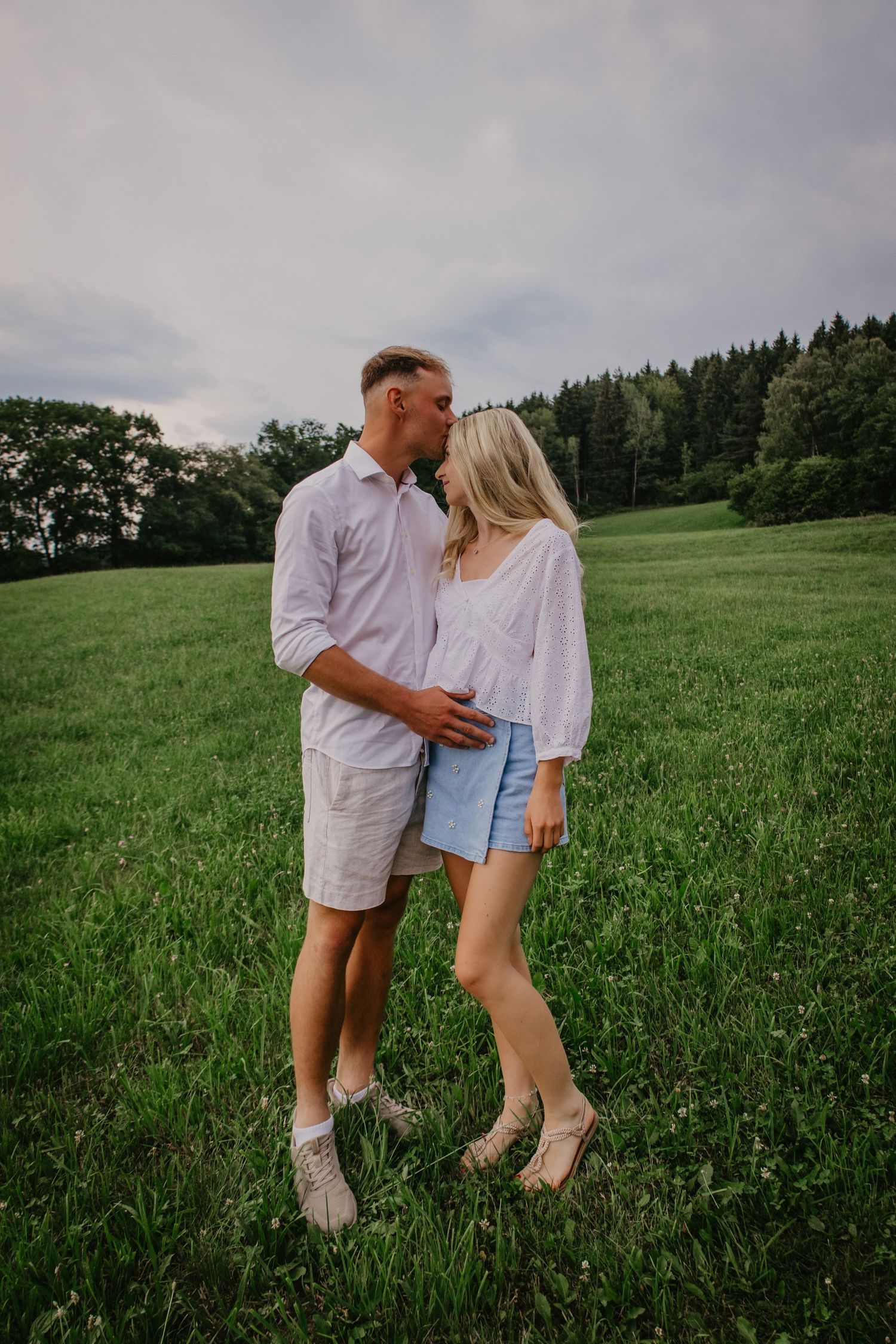 A couple sharing a kiss in a lush green meadow, dressed in casual white and blue summer outfits.