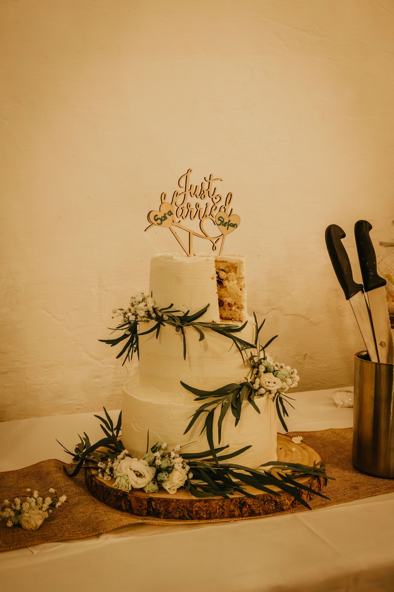 Three-tier white wedding cake with greenery, flowers, and a Just Married topper on a wooden slice.
