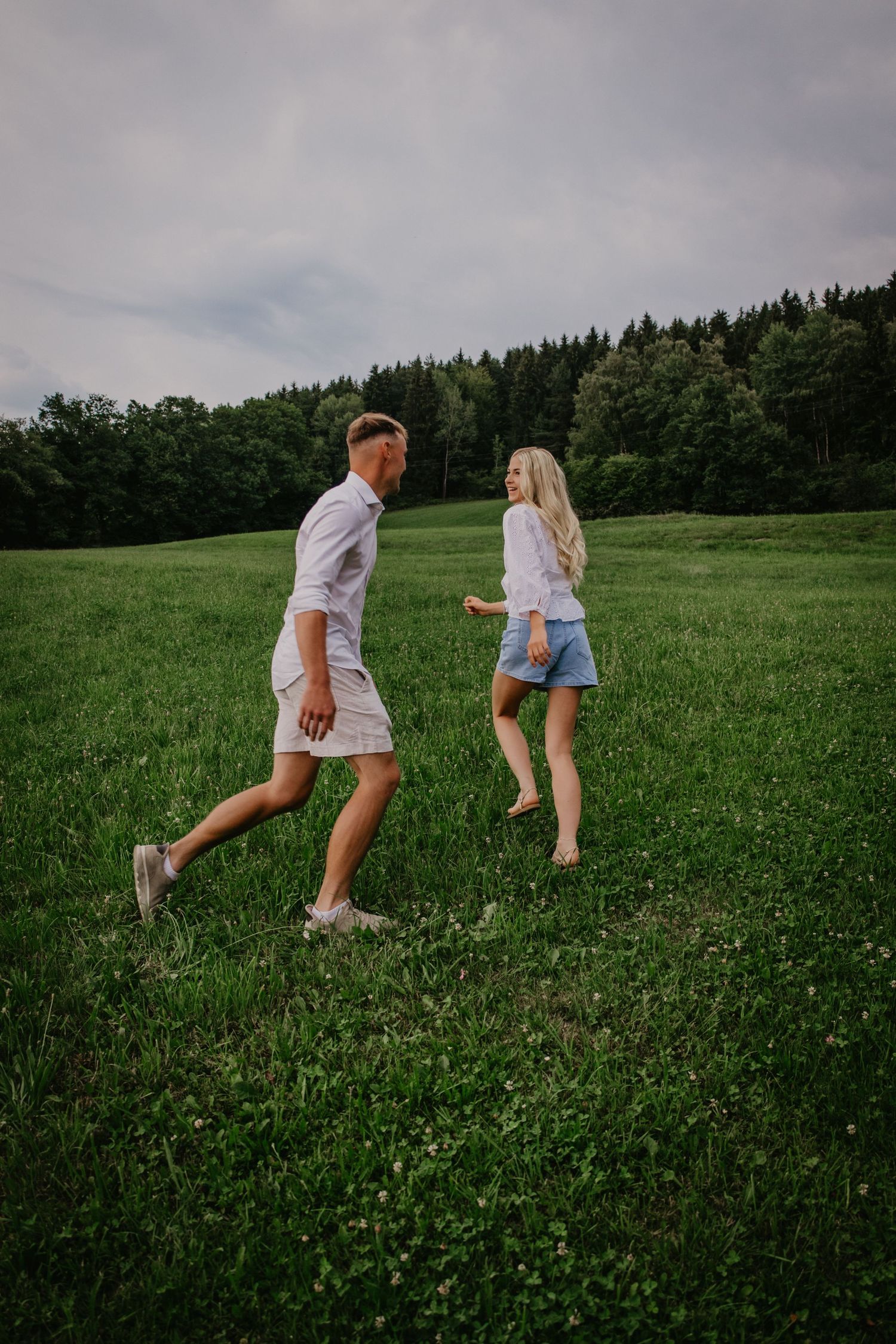 A young couple walks barefoot through a lush green meadow with a forest backdrop on a cloudy day.