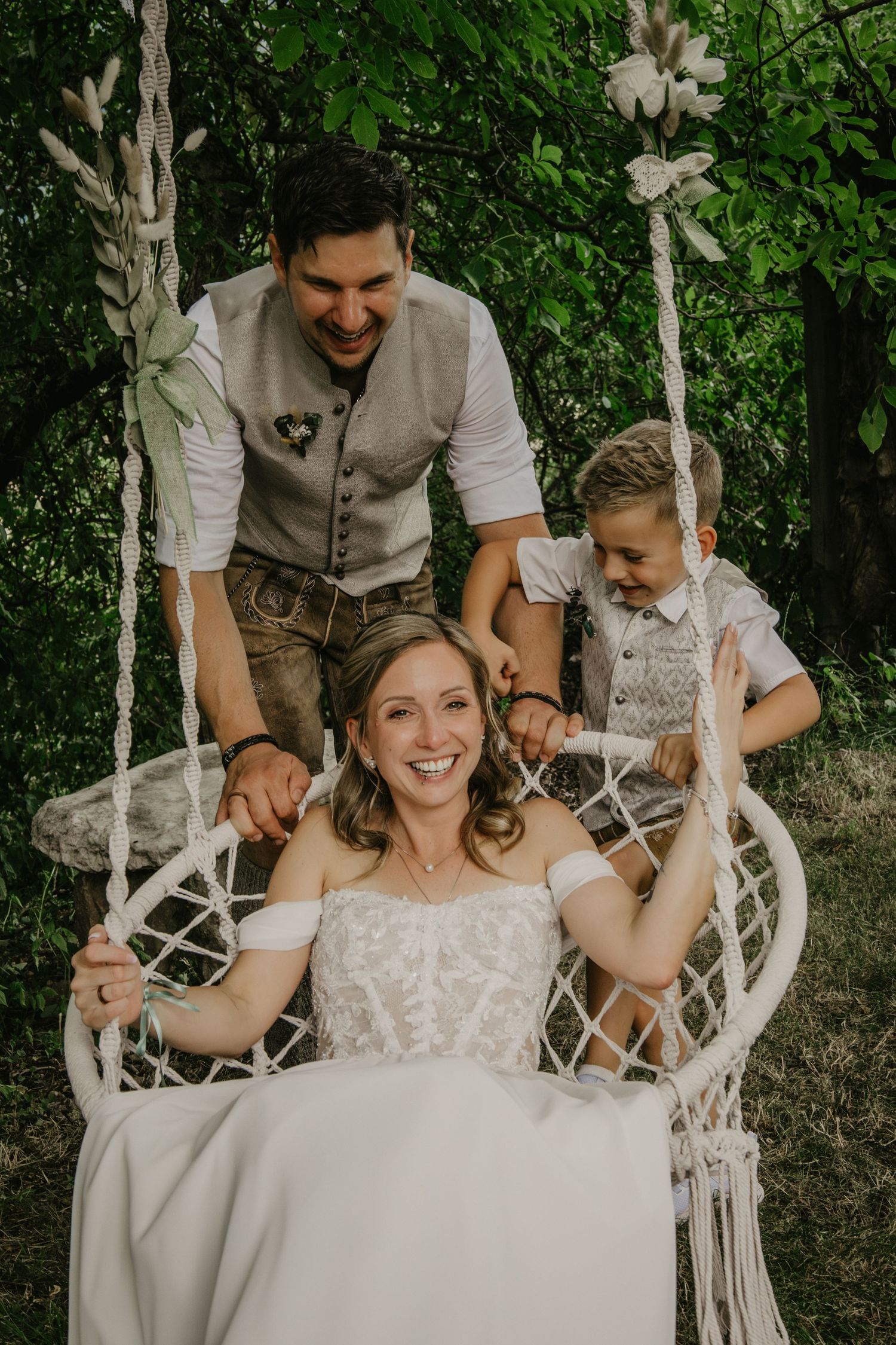 Bride laughing on macrame swing while groom and boy push her in lush garden wedding setting.