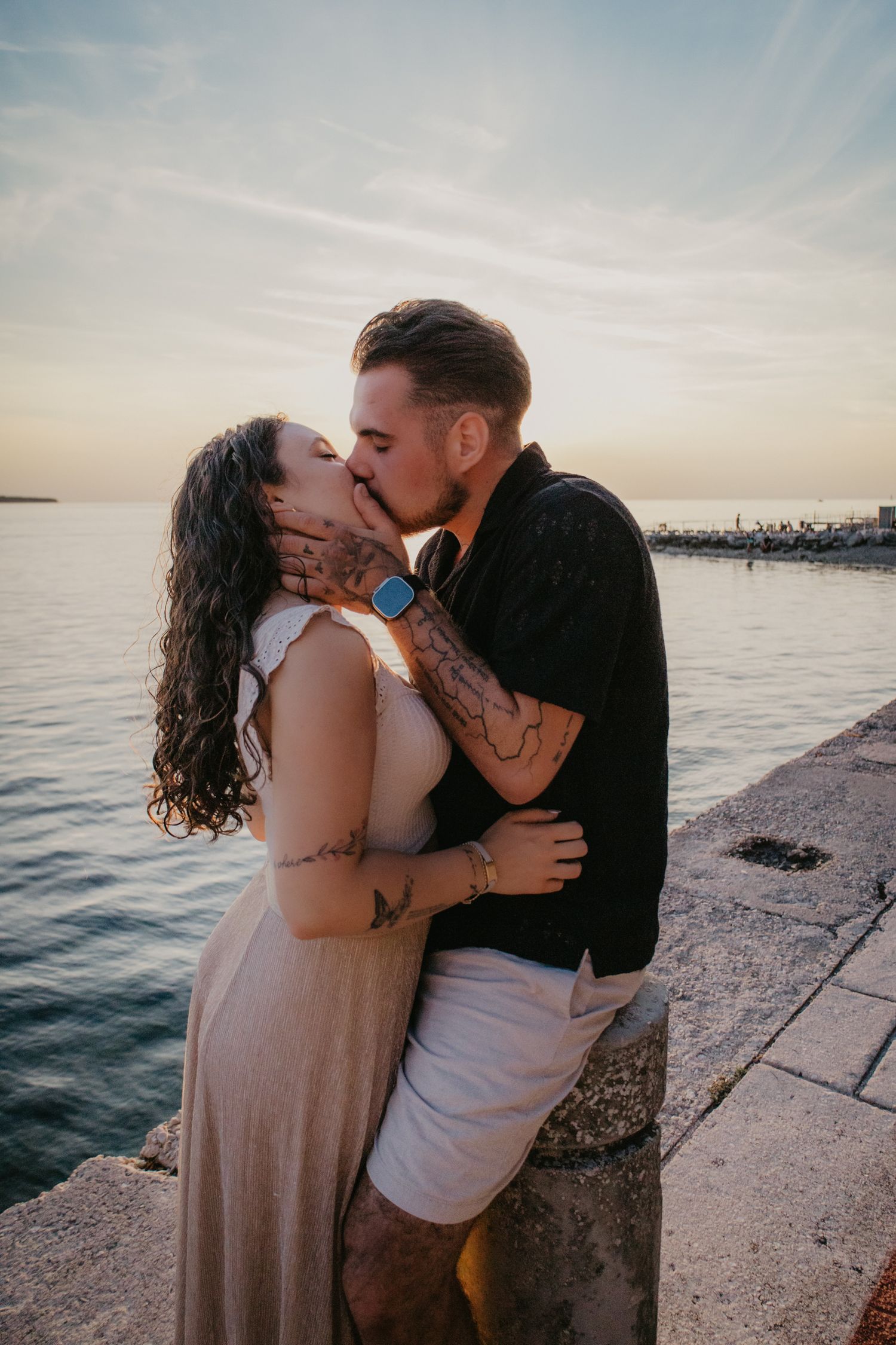 A romantic couple embraces on a concrete pier with the ocean and sunset behind them.
