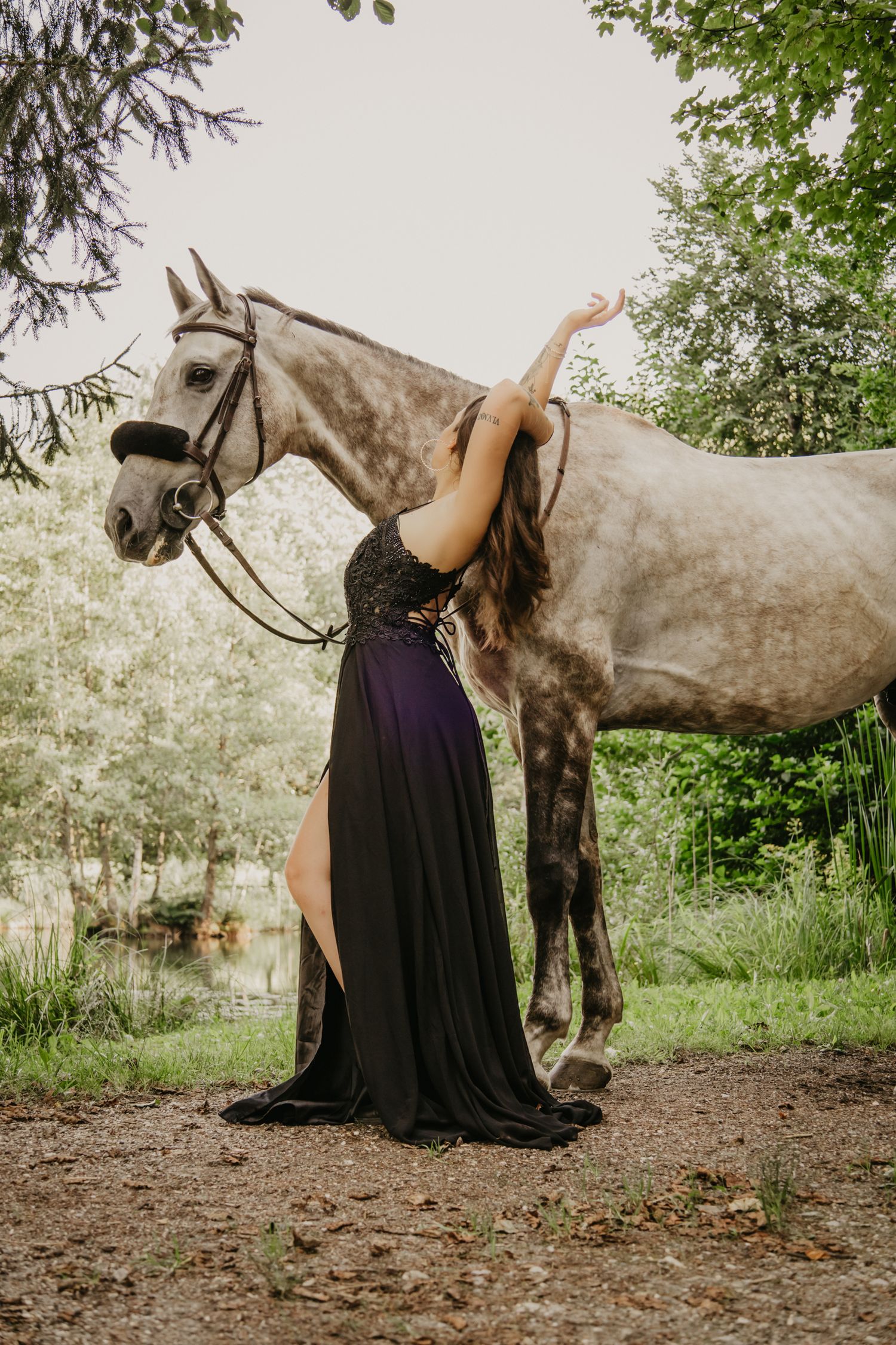 An elegant dance-like pose in a flowing black dress beside a grey horse in nature.