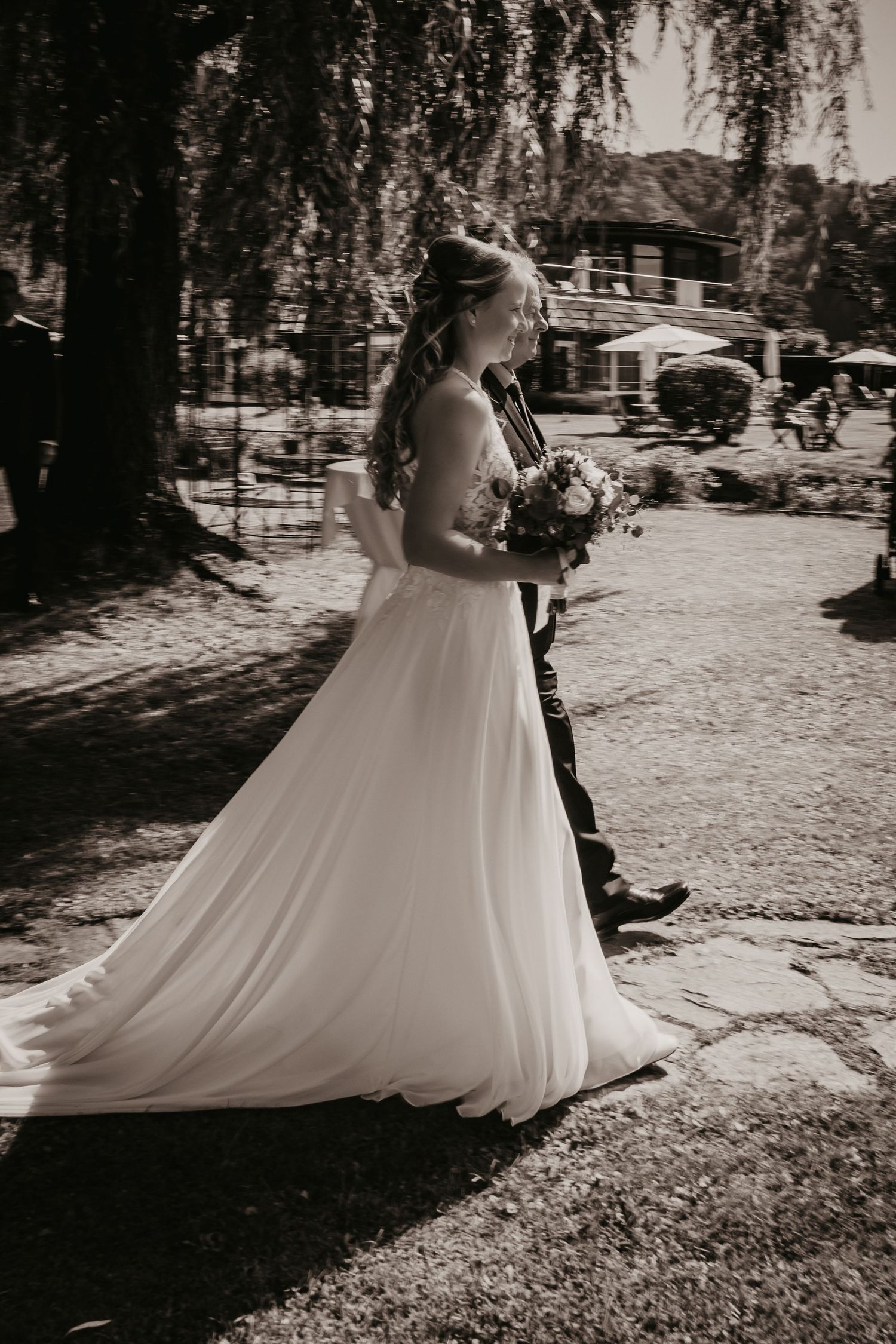 A flowing white wedding dress train trails across a sunlit garden path.