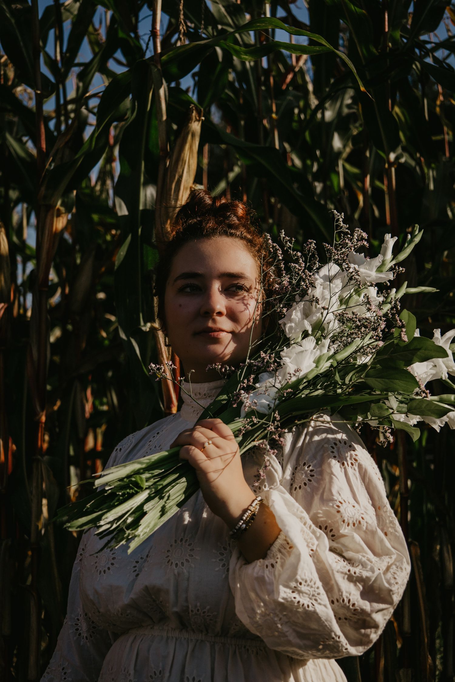 Artistic portrait with white flowers and vintage dress among corn stalks at sunset.