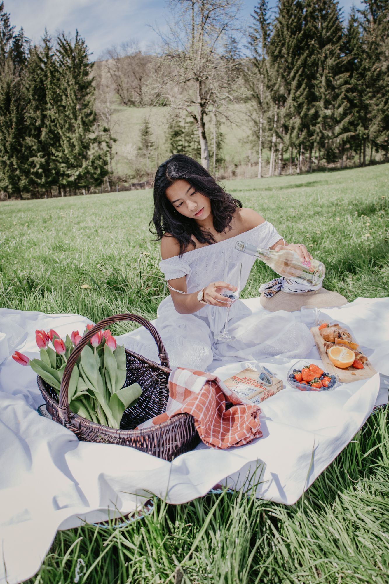 Spring picnic setup on white blanket with fresh food and flowers in grassy meadow.
