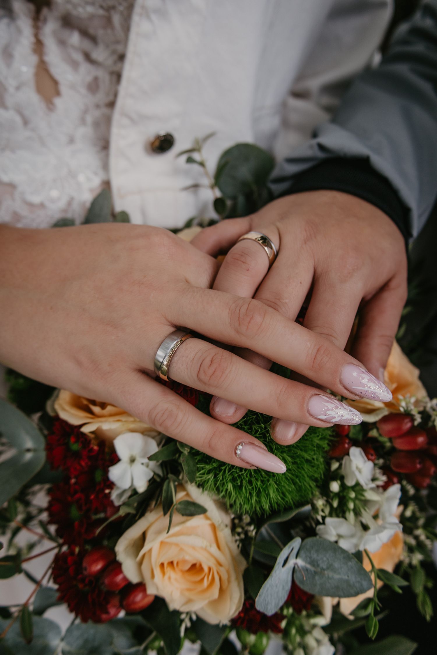 Close-up of hands with wedding rings touching a peach and burgundy floral bouquet.