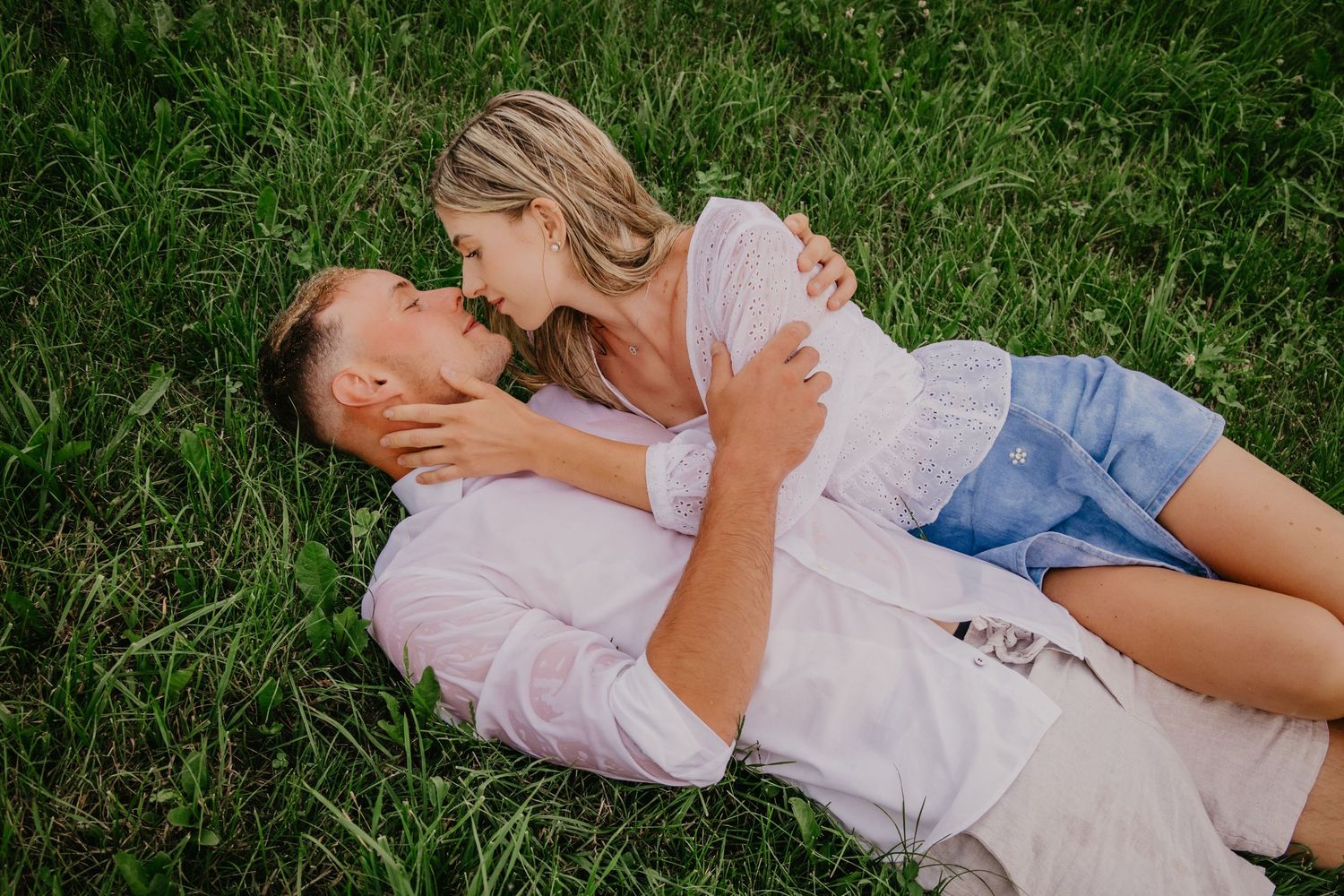 A romantic couple lying in green grass, woman cradling man's face as they share an intimate moment outdoors.