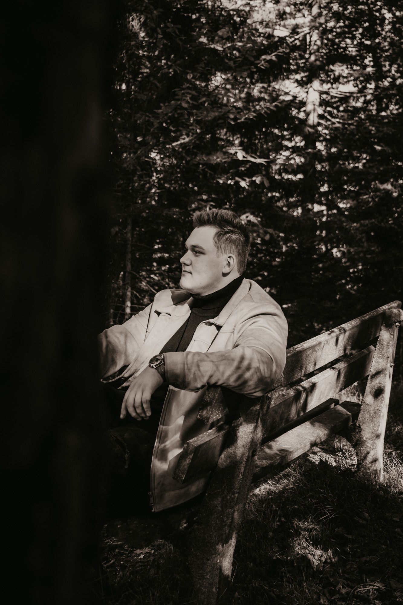 Moody black and white portrait of someone resting against a wooden fence in a forest setting.