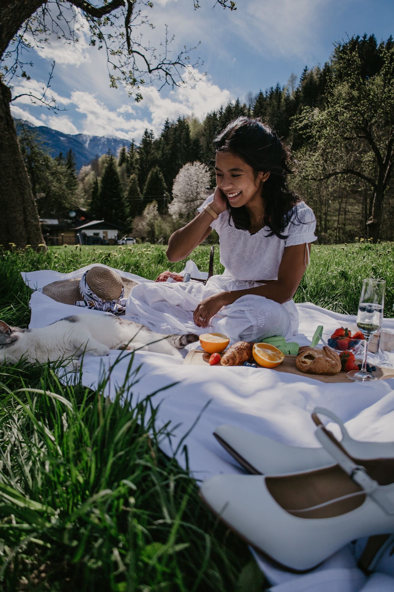 A summer picnic setup with fresh fruits and snacks laid out on a white blanket in a mountain meadow.