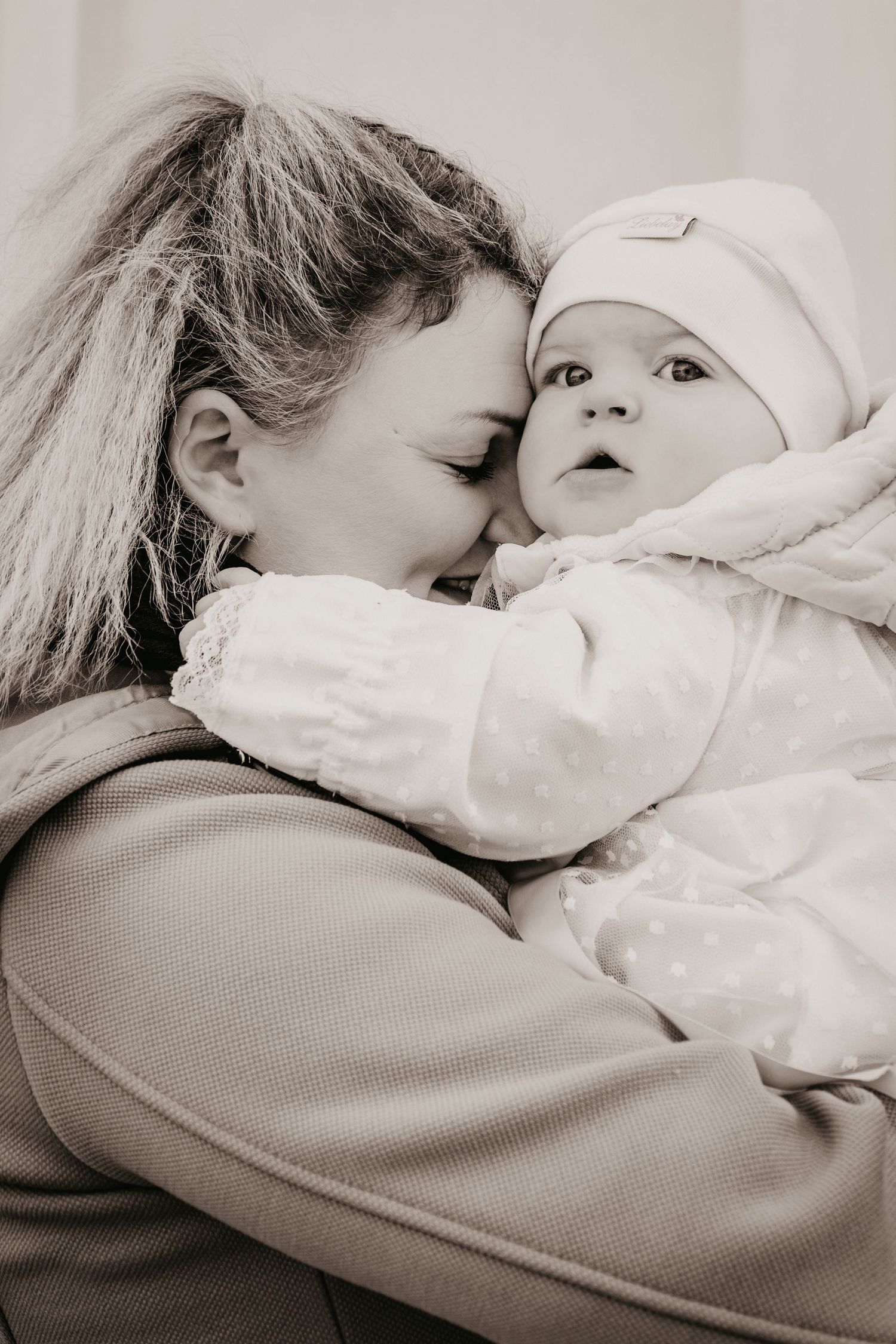 A woman with long hair tenderly hugs a baby in a white hat and polka dot snowsuit in a warm sepia-toned portrait.