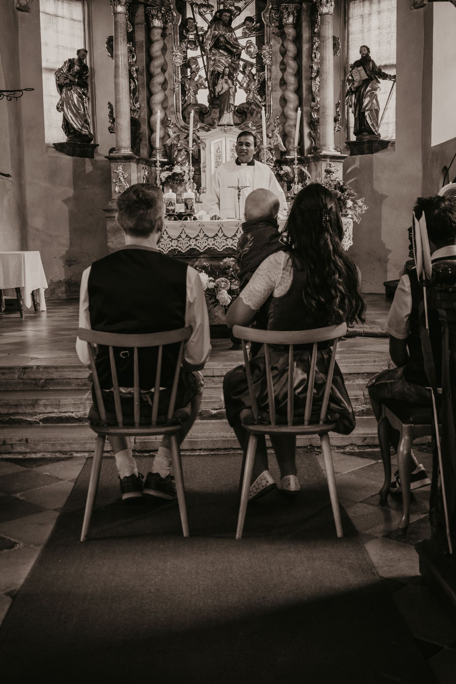 Couple in traditional attire sit on wooden chairs facing a priest at an ornate baroque church altar during a wedding ceremony.