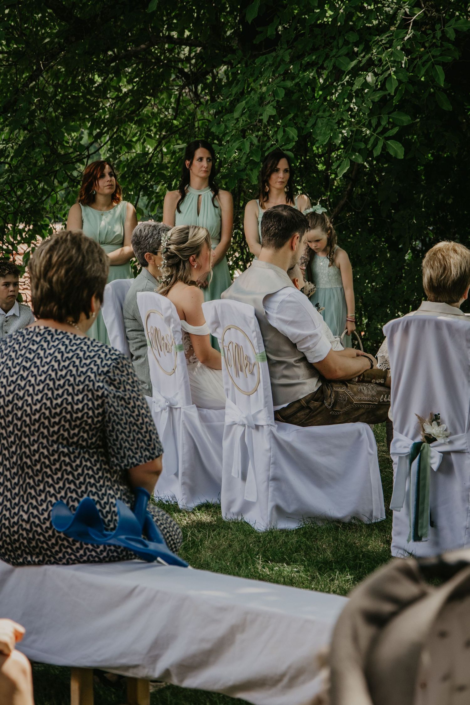 Outdoor wedding ceremony with bride and groom seated in white chairs, bridesmaids in mint dresses standing beneath green trees.