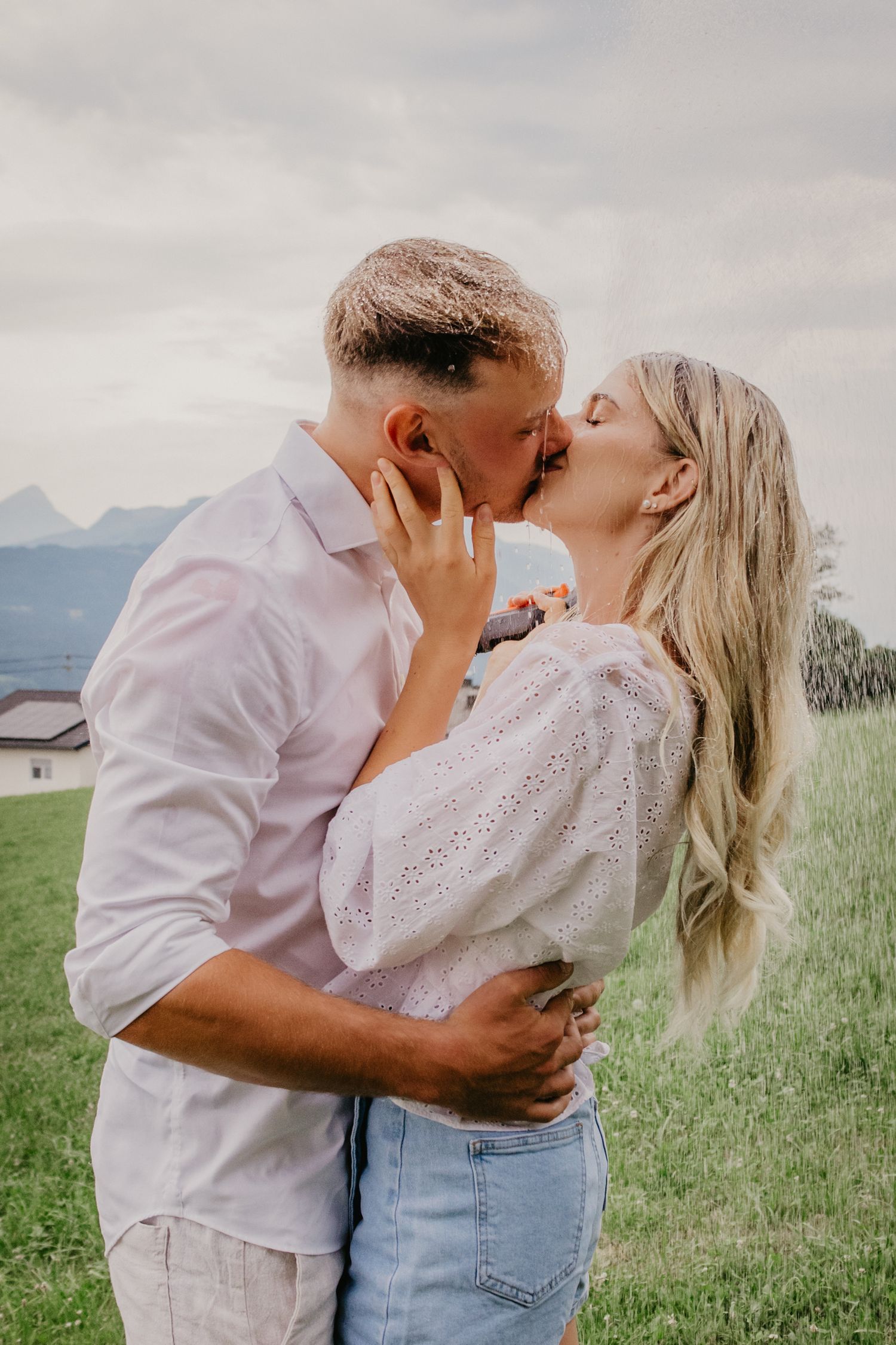A couple shares a romantic kiss outdoors on a green hillside with mountain views, the woman in white top and jeans.