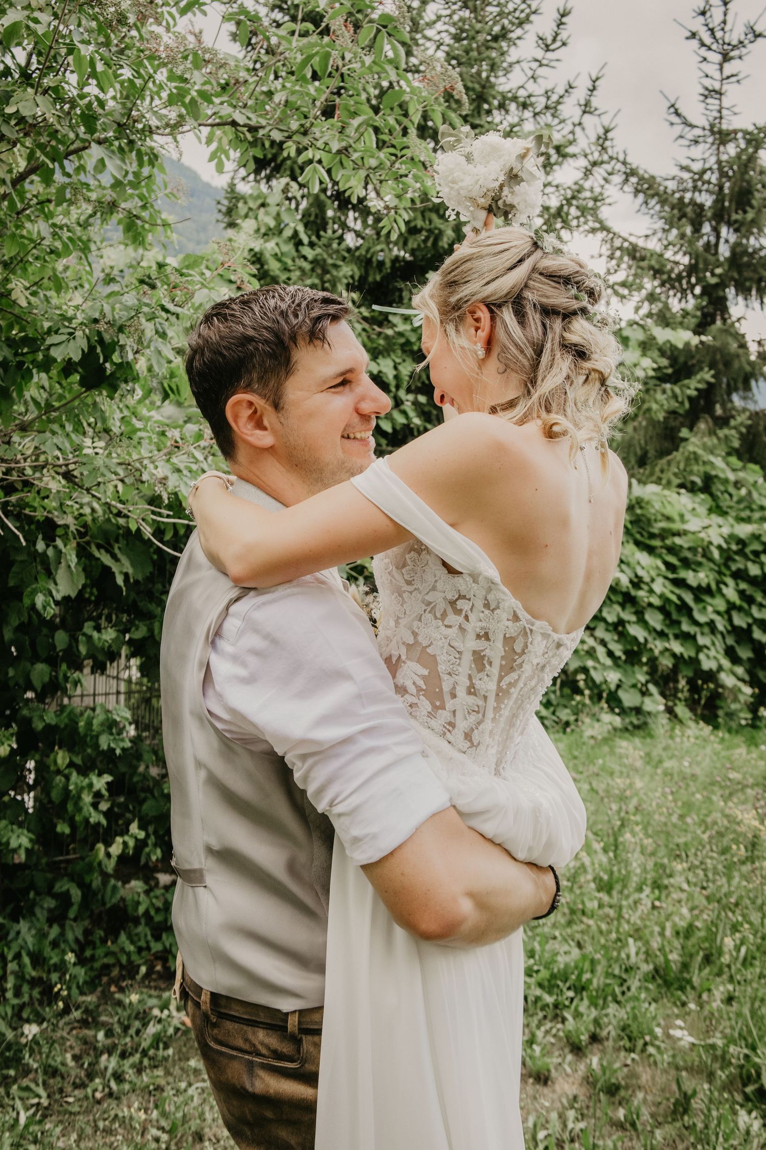 Groom smiling while embracing bride in lace off-shoulder wedding dress outdoors in green garden.