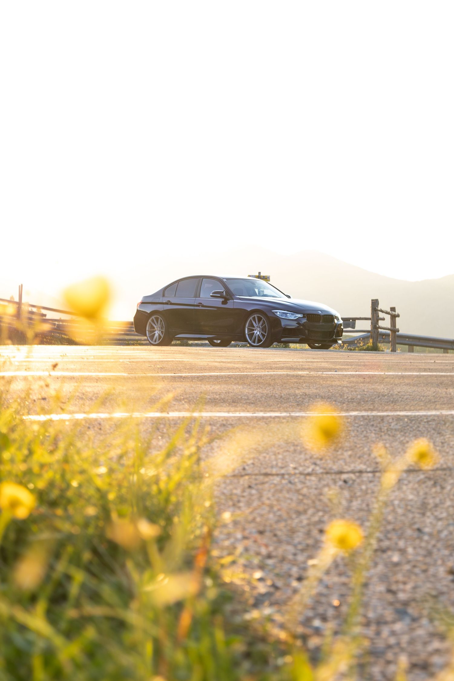 Black BMW sedan parked on a mountain road at golden sunset, framed by yellow wildflowers in the foreground.
