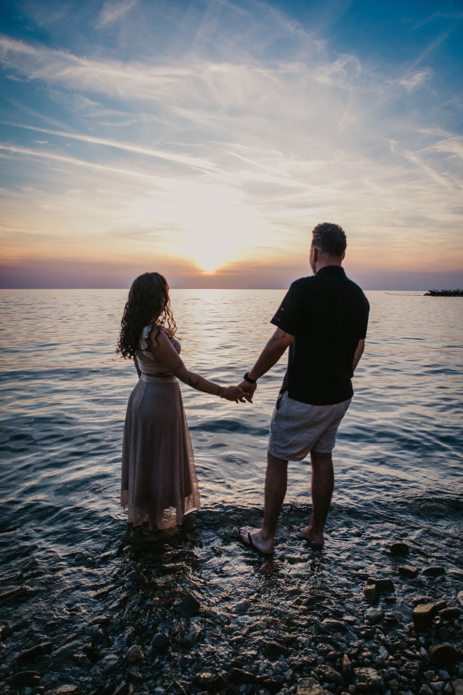 Couple standing in shallow ocean water at sunset creating silhouettes against the colorful sky.