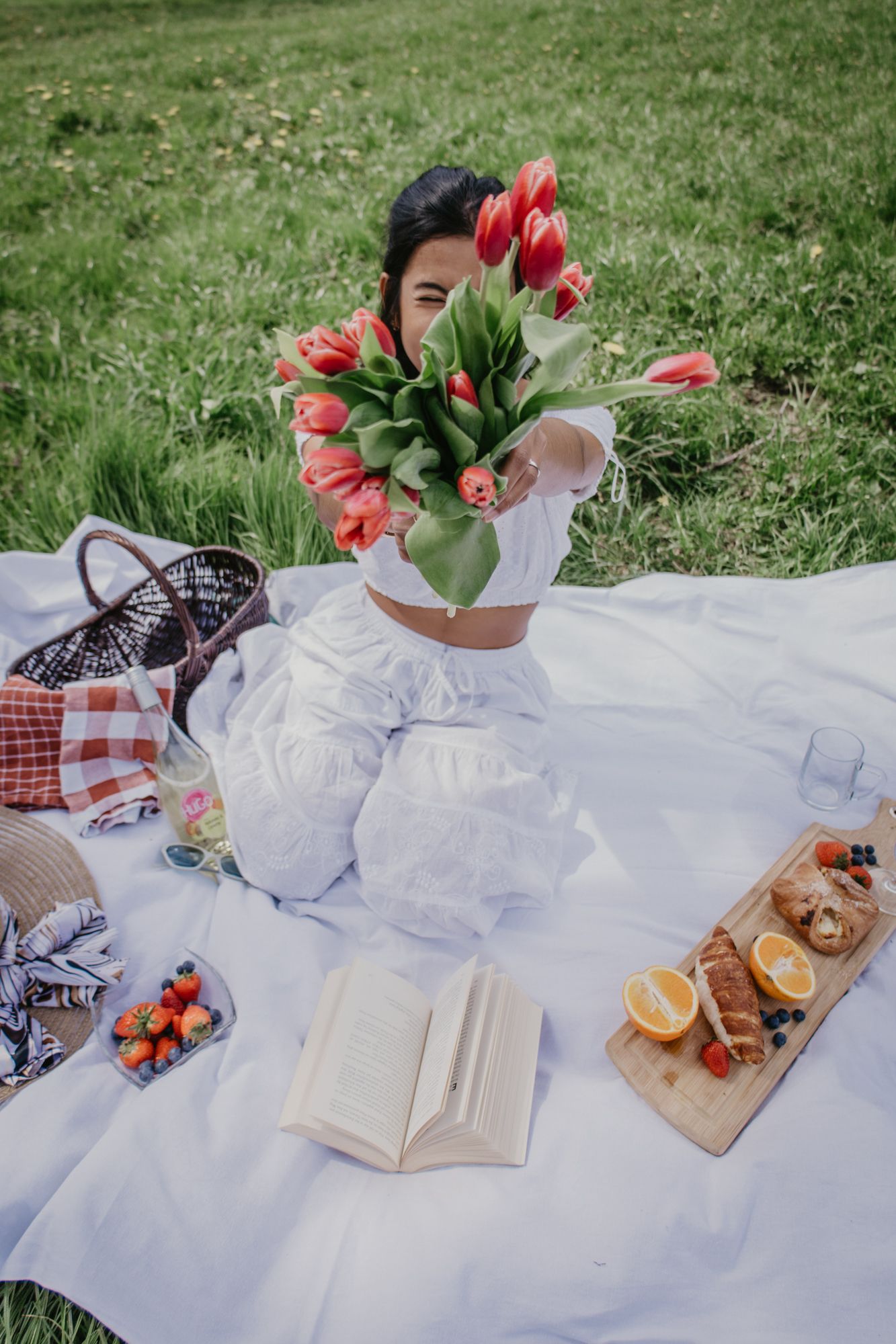 An elegant picnic display with red tulips, books, and refreshments on a white blanket outdoors.