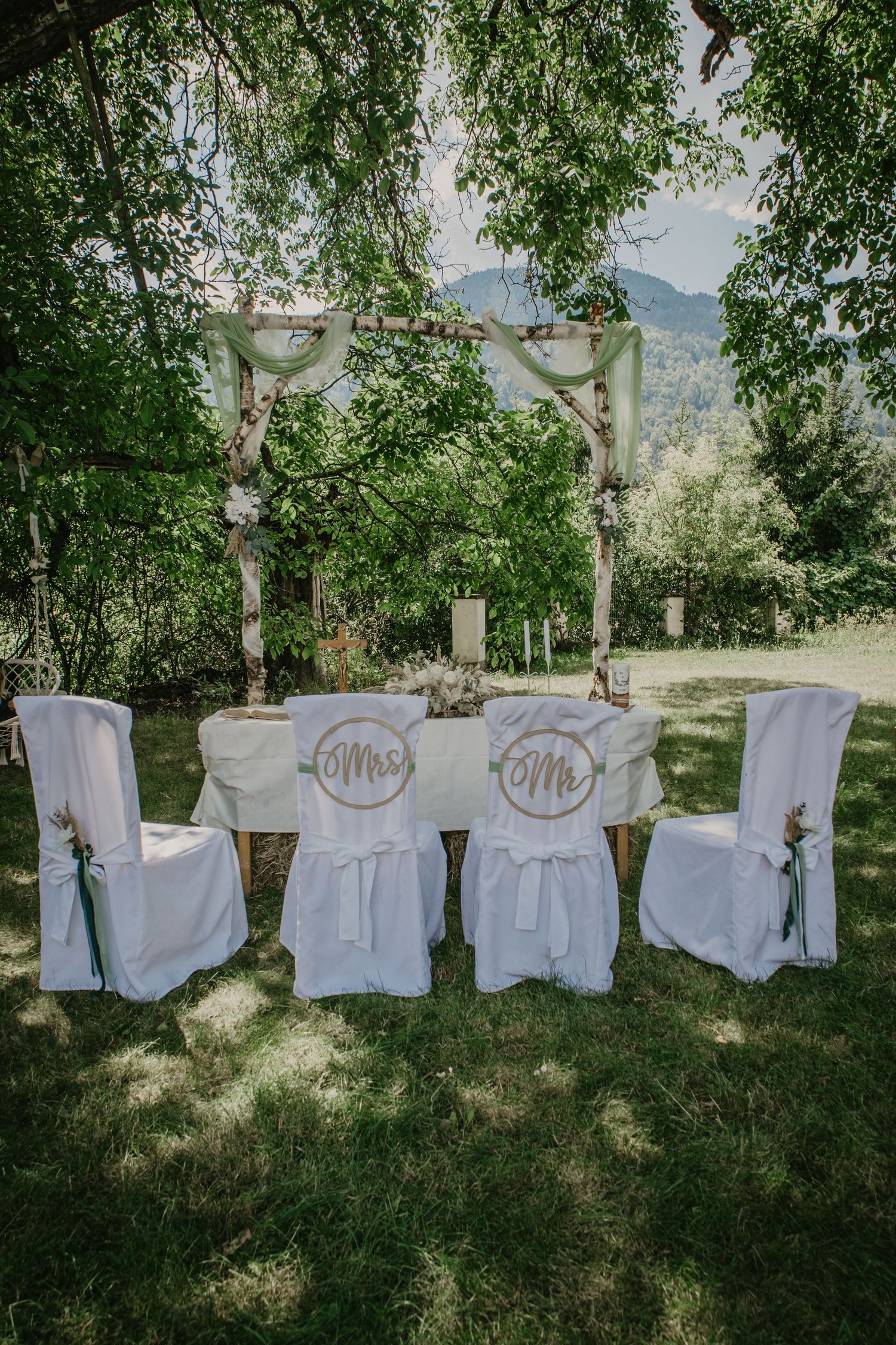 Outdoor wedding setup with white chair covers labeled Mrs and Mr beneath a rustic birch arch surrounded by lush greenery.