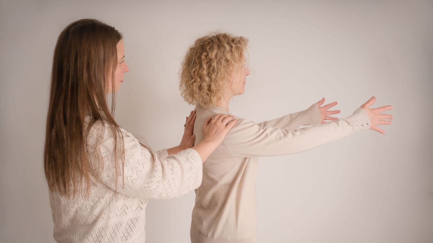 Two people in light colored clothing stand side by side with arms outstretched against a neutral background doing somatic coaching session.