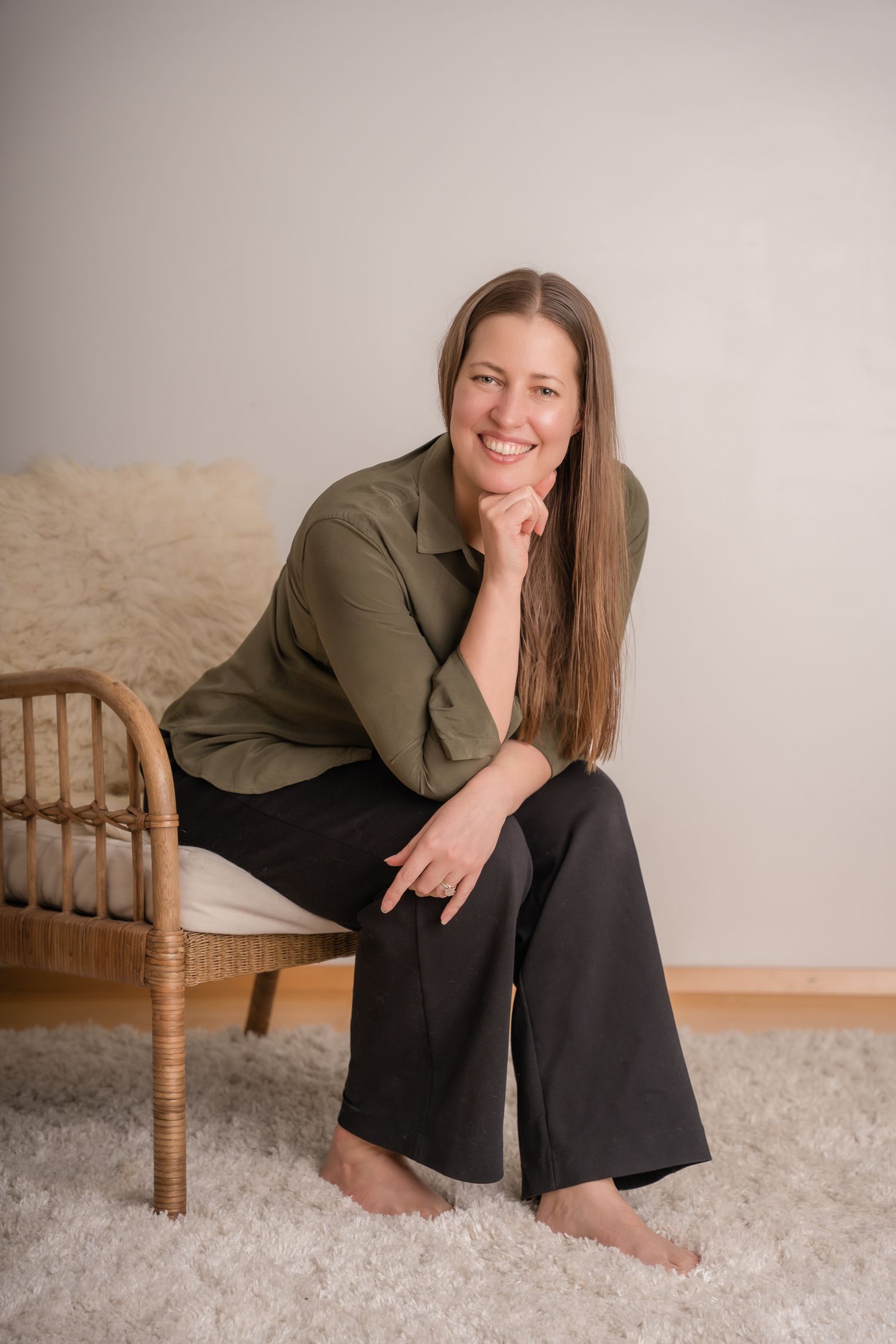 A person in an olive green shirt and black pants sits casually on a wooden chair against a neutral background.