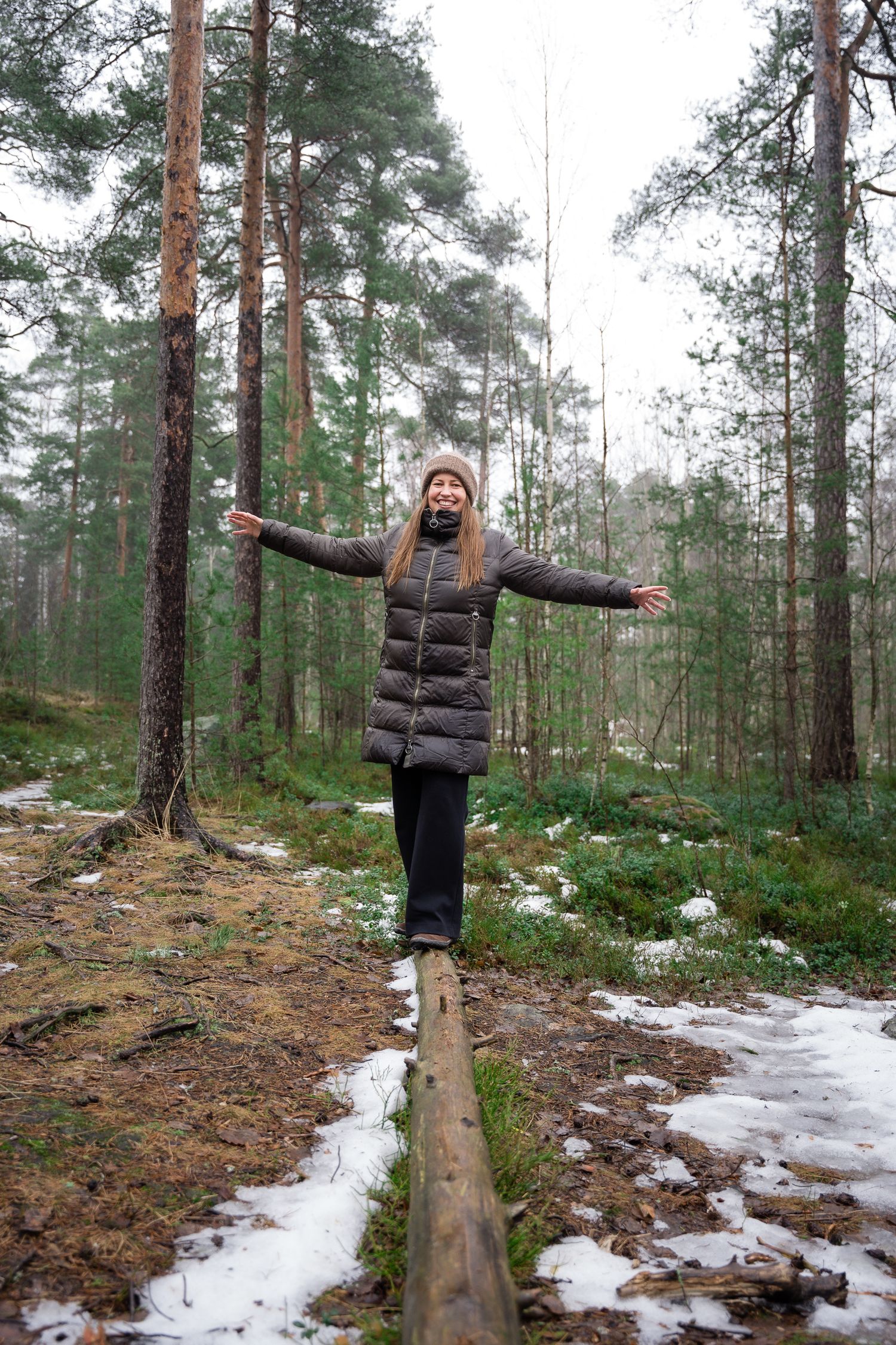 Woman balancing on a log in a snowy winter forest, arms outstretched, wearing a dark puffer jacket and beanie.