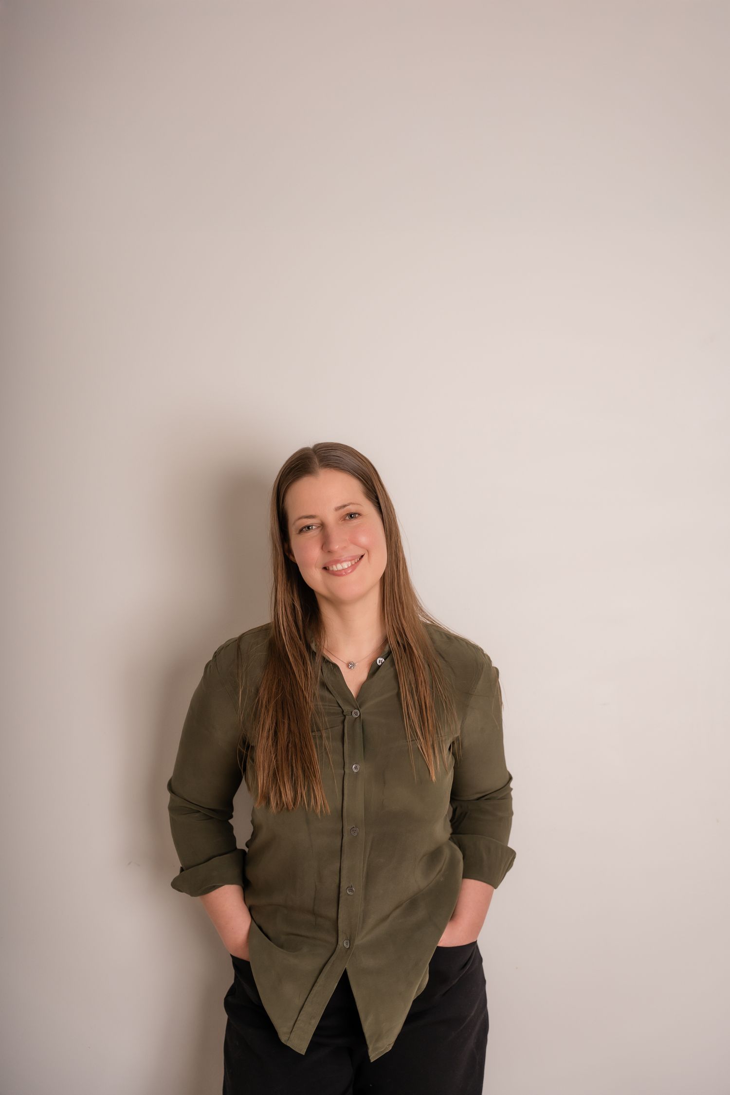 Smiling woman with long brown hair wearing an olive green button-up shirt, standing against a light gray wall.