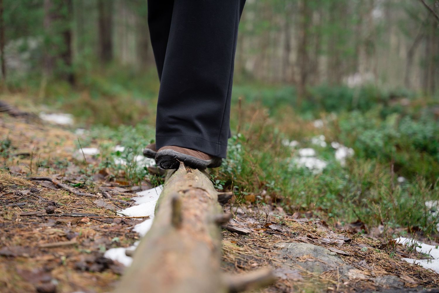Person balancing on a fallen log in a forest trail wearing black pants and brown shoes.