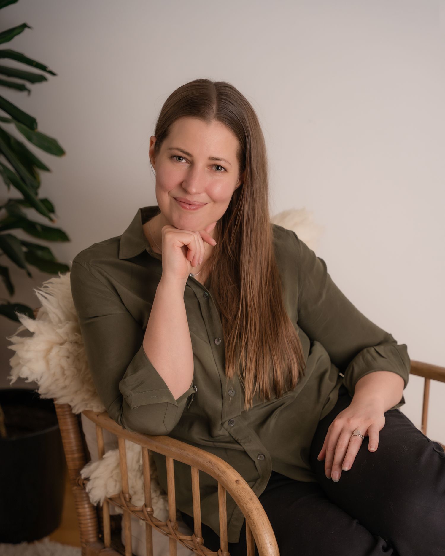 A woman in an olive green top lounges casually on a wooden rocking chair against a neutral wall with indoor plants.