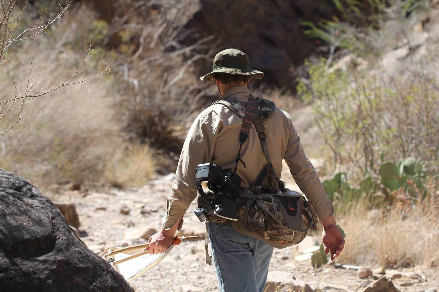 A person hiking along a rocky desert trail wearing outdoor gear and a backpack, looking for jumping spiders.