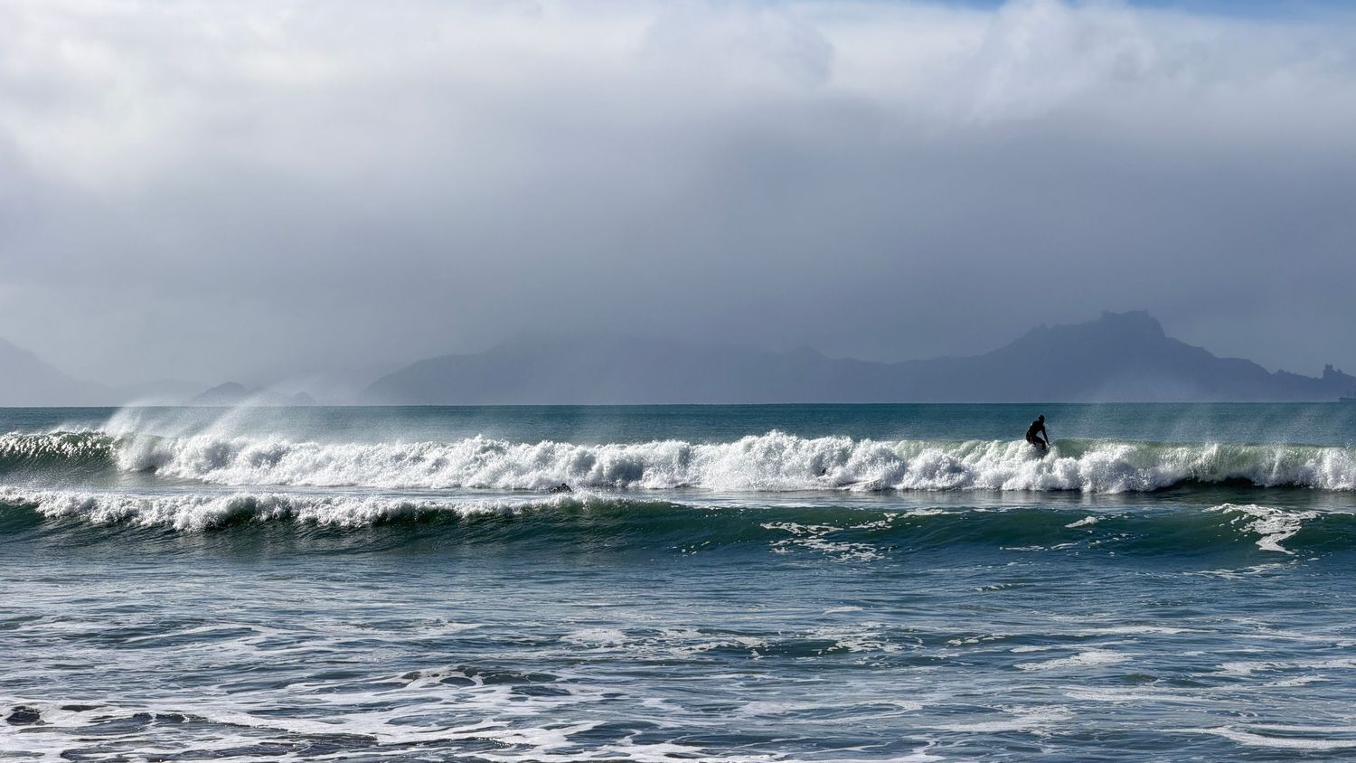 Surfer riding ocean waves with dramatic cloudy skies and mountain silhouette in the background.