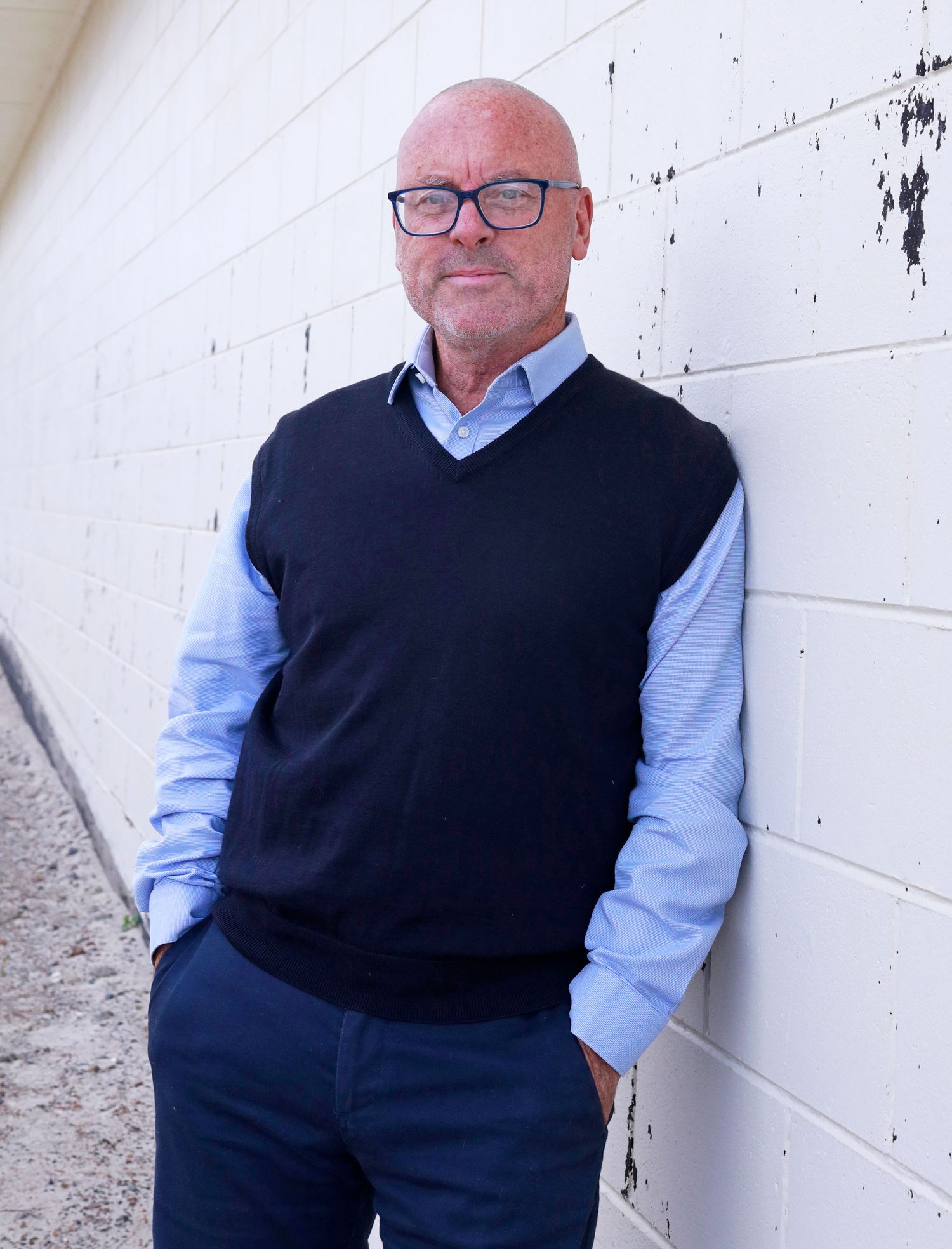 Bald man with glasses wearing a navy vest over a light blue shirt, leaning against a white brick wall.