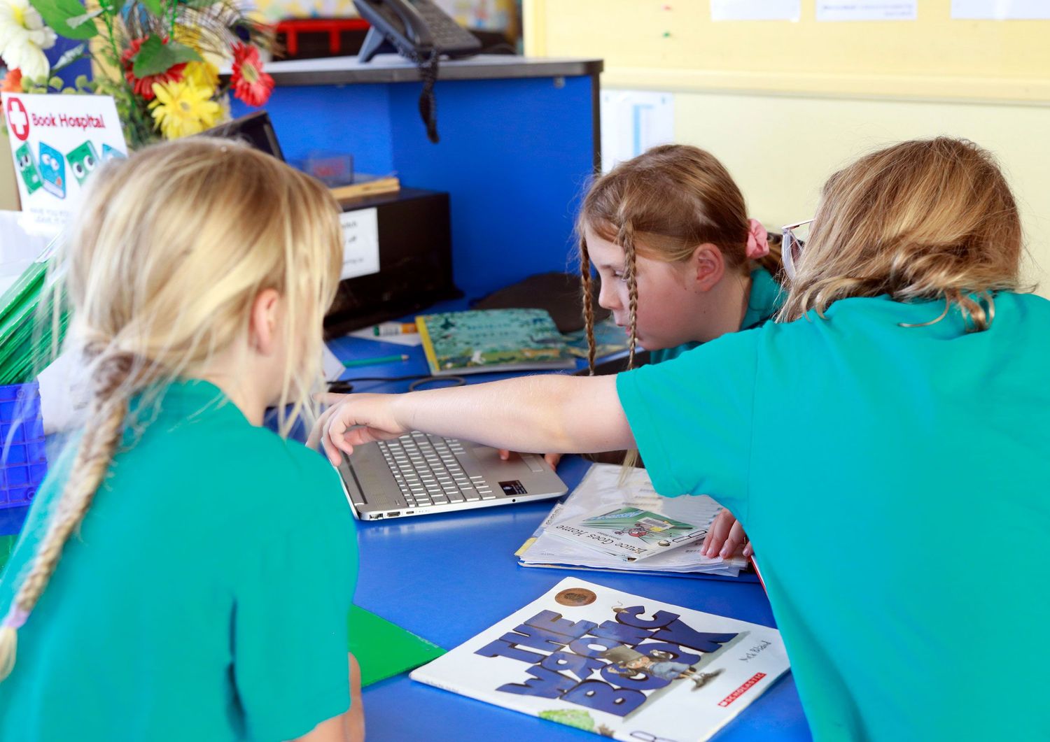 Three children in green shirts collaborate on a laptop computer during a classroom learning activity.