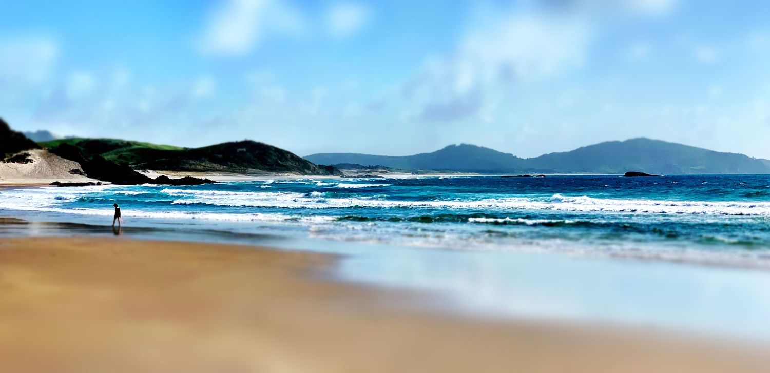 A lone figure walks along a pristine sandy beach with turquoise waves and green hills under a blue sky.