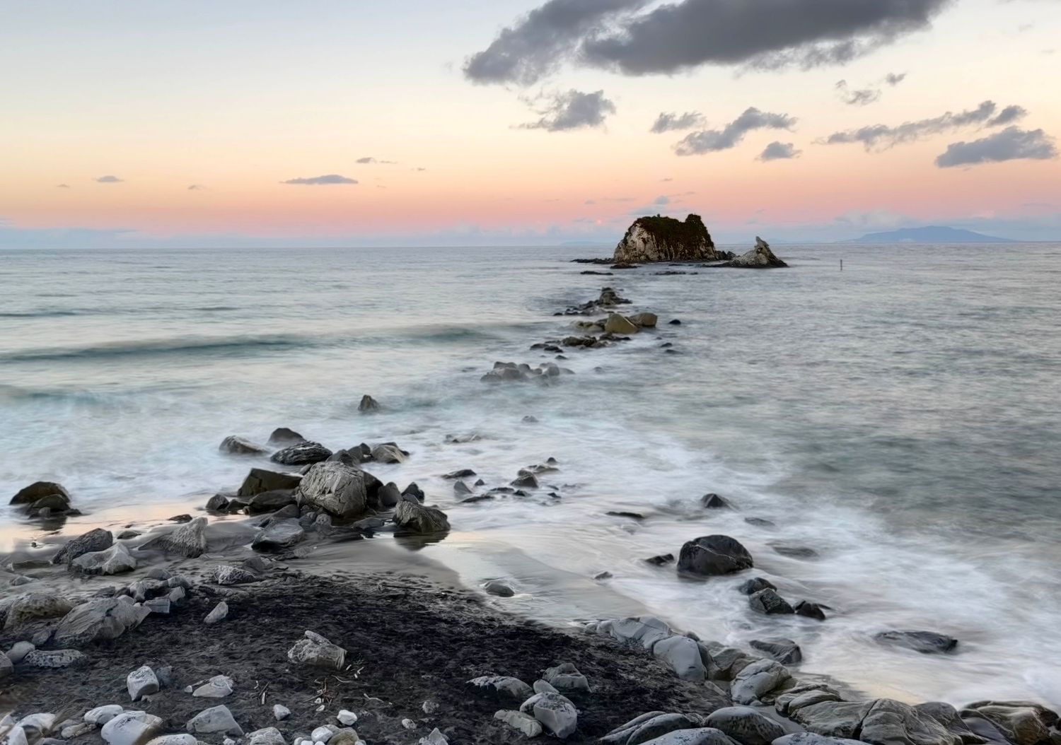 Rocky shoreline at sunset with misty ocean waves and a small island silhouette against a pastel pink and purple sky.