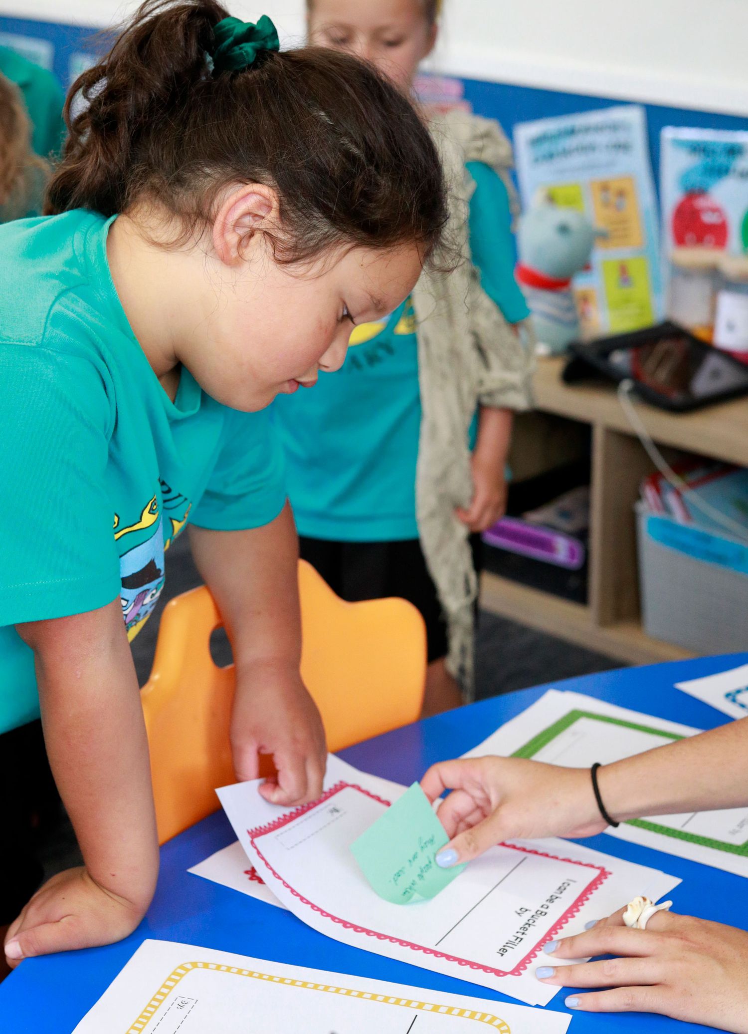 Young girl in teal shirt working on a worksheet activity at a blue table in a colorful classroom.