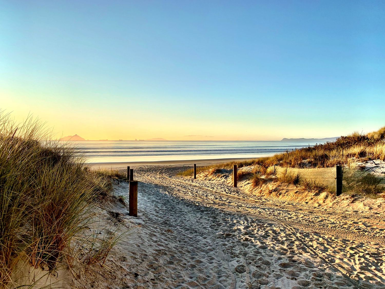Sandy beach path with wooden posts and dune grass leading to a calm ocean at golden sunset.