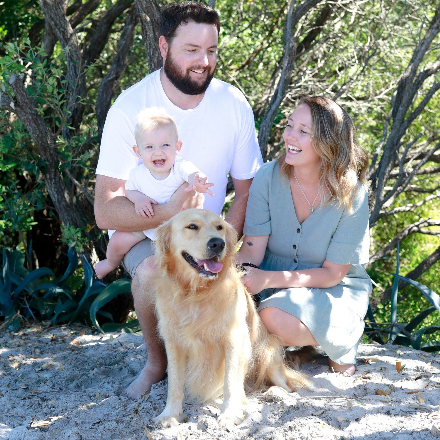 A happy family with a baby, golden retriever dog posing on sandy beach with lush green trees in background.