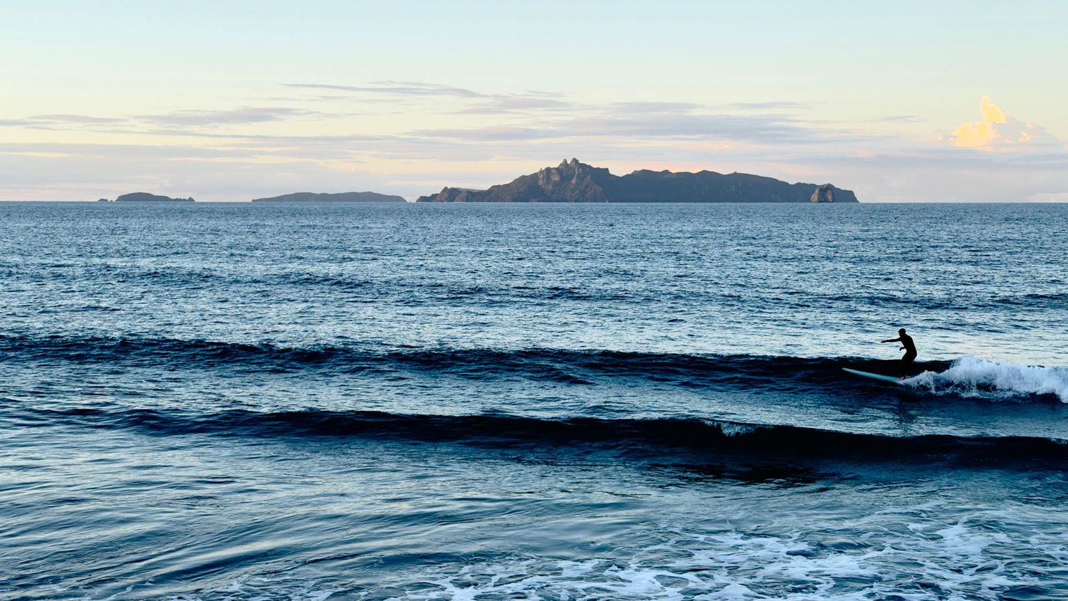 Surfer riding ocean waves at dusk with a small island visible in the calm blue sea background.