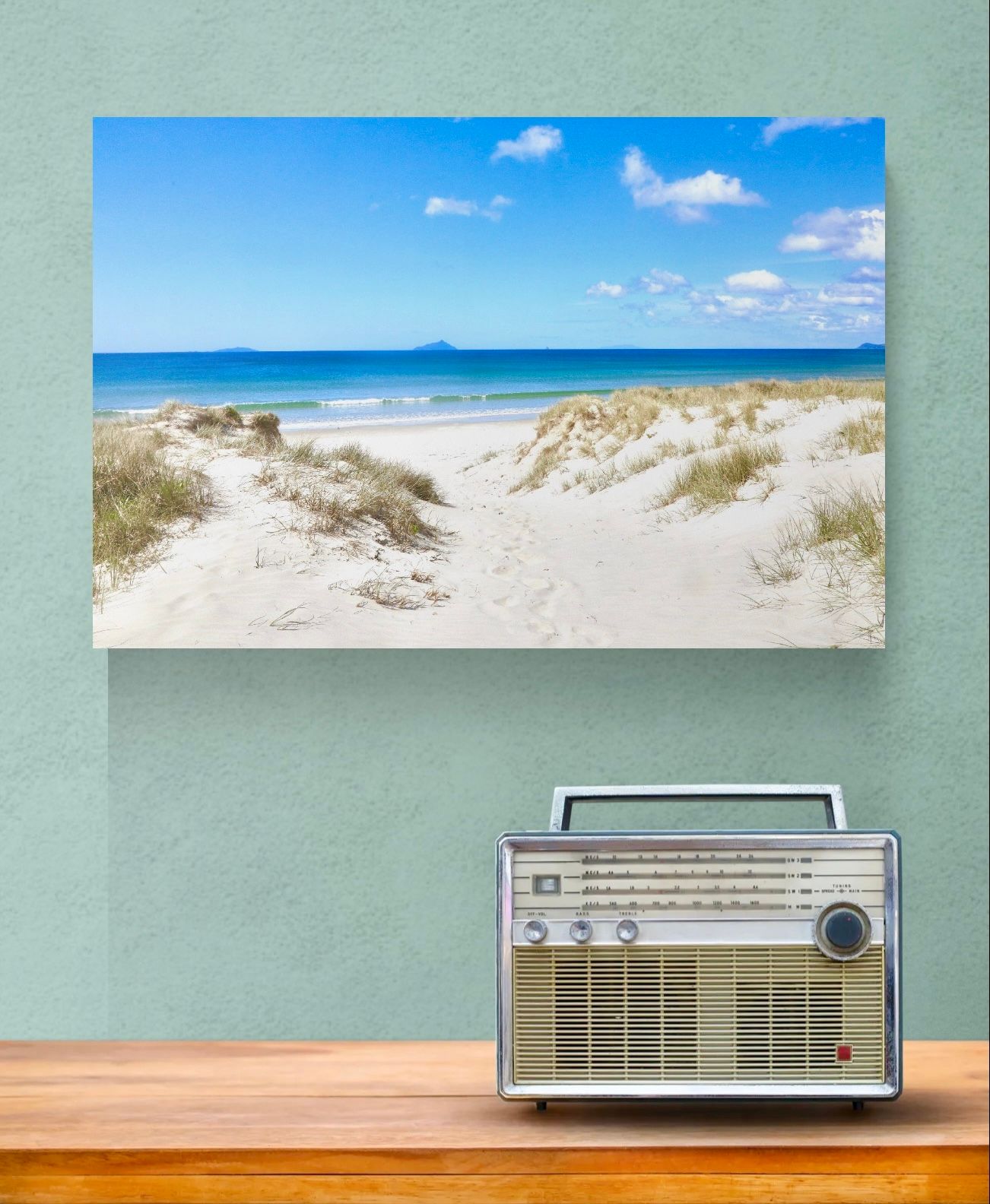 Vintage radio on wooden shelf below a canvas print of a sunny beach with white sand dunes and blue sky.