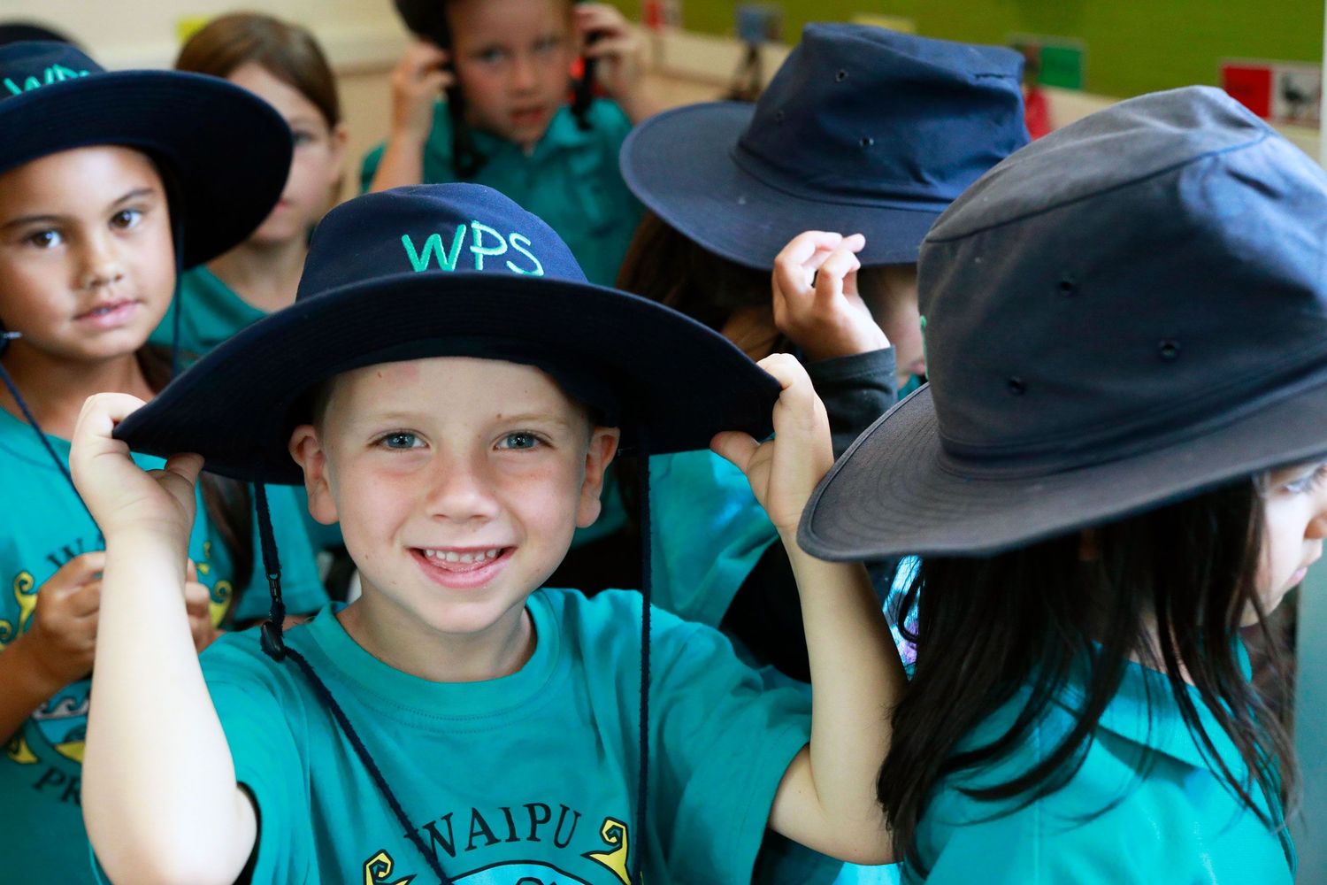 Children wearing teal WPS school uniforms and wide-brim hats smile outdoors during a school event.