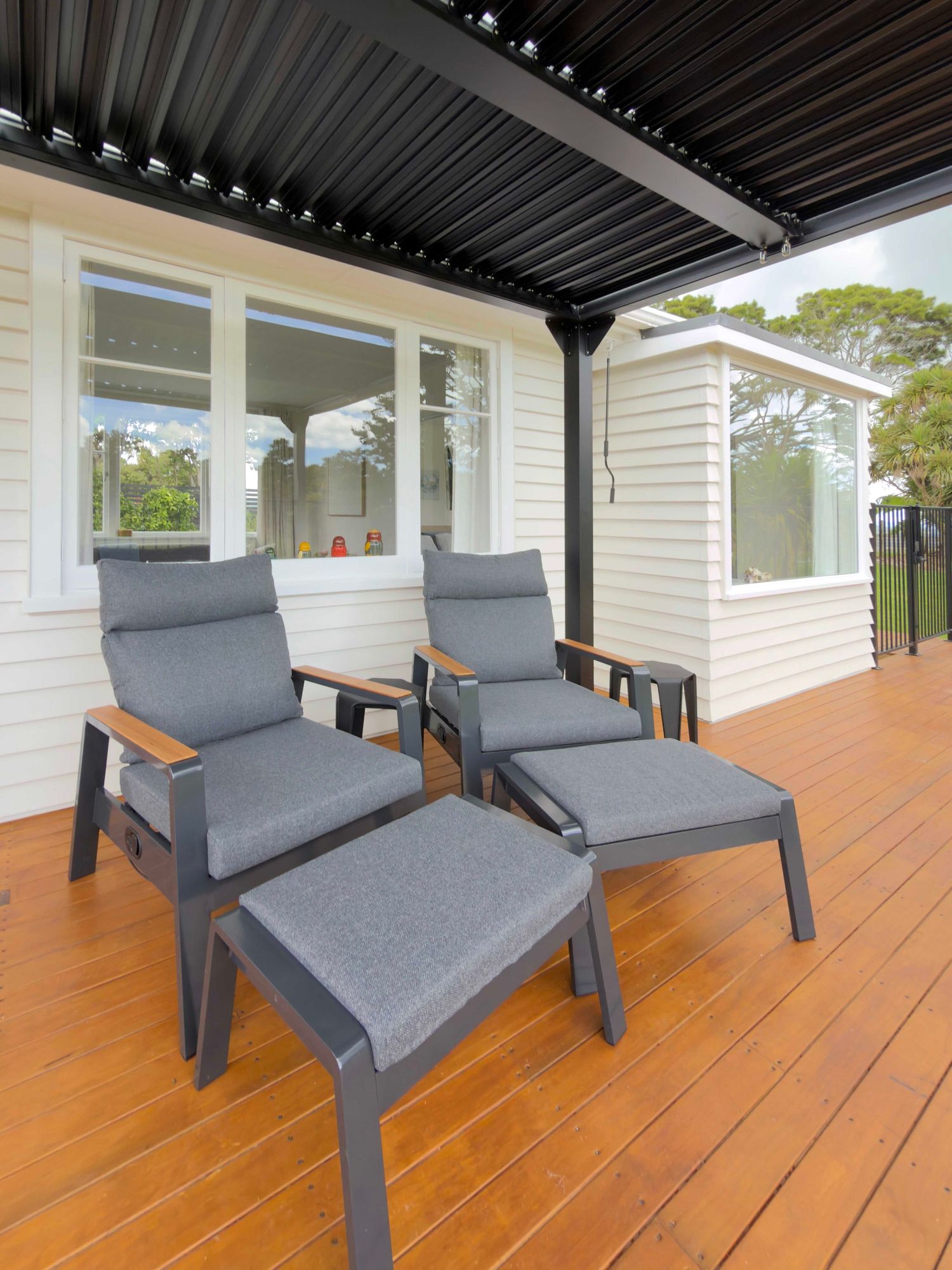 Modern outdoor deck with four grey cushioned lounge chairs and ottomans under a black pergola beside a white house.