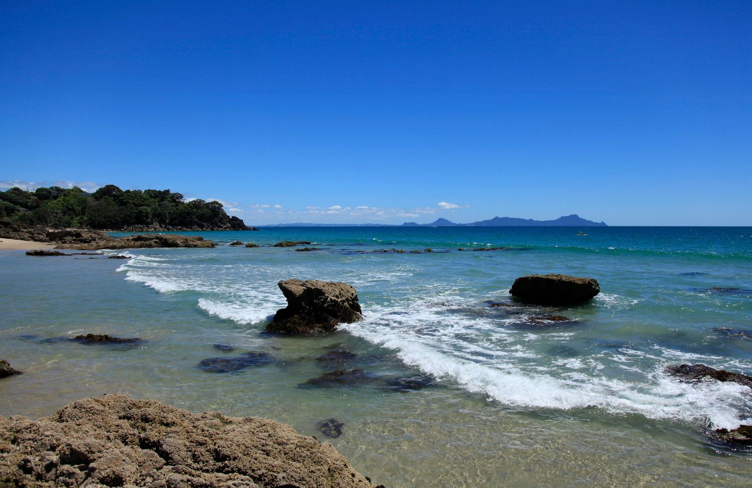 Rocky shoreline with clear turquoise water, gentle waves, and a stunning blue sky over a distant island coastline.