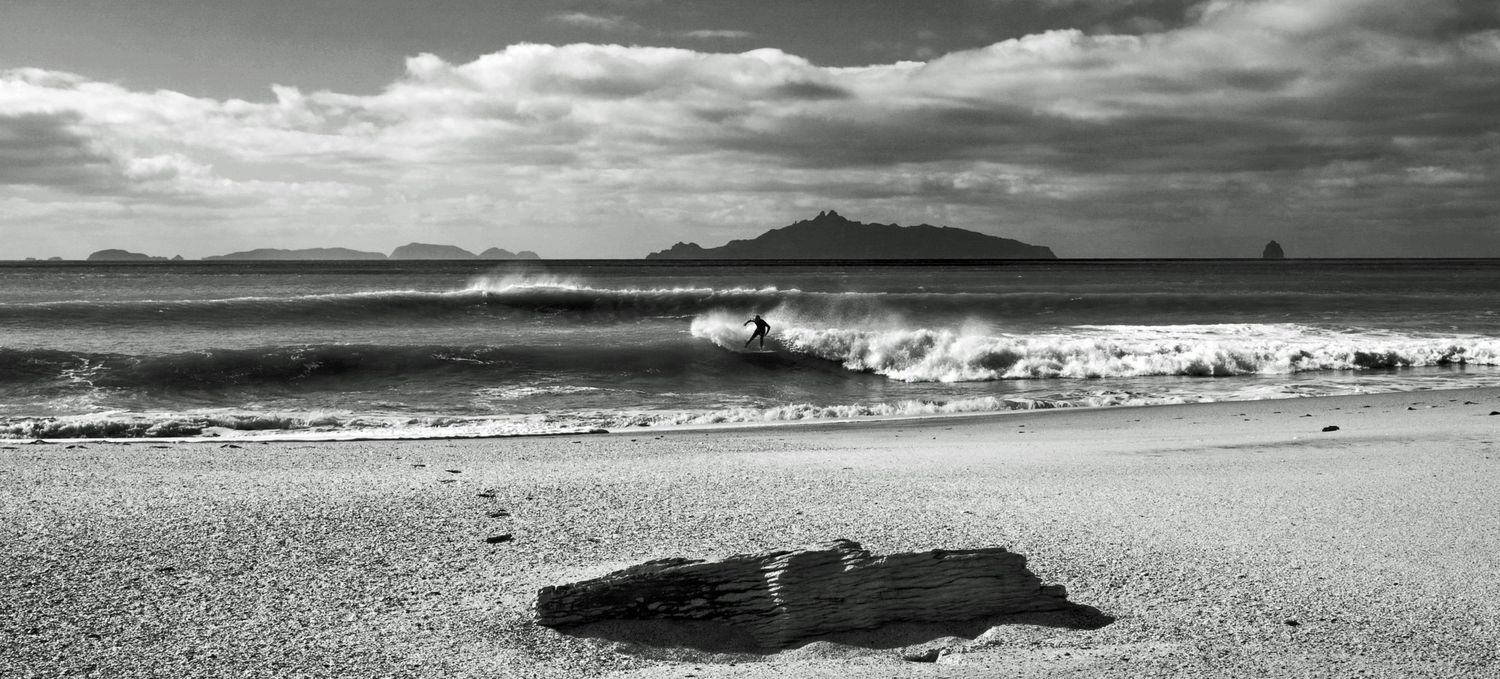 Black and white coastal beach scene with ocean waves, driftwood foreground, and distant island mountains under dramatic cloudy sky.