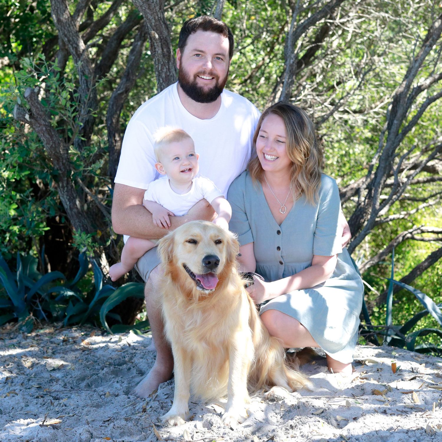 Family with baby and golden retriever dog posing on sandy beach surrounded by lush green trees on a sunny day.