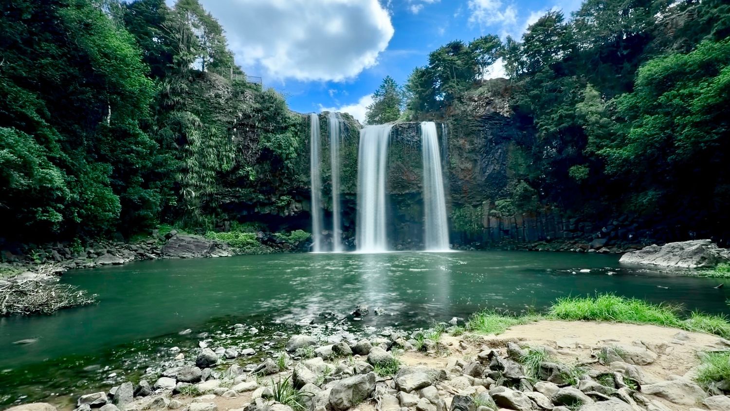 Majestic waterfall cascading into a serene rocky pool surrounded by lush green tropical forest under a partly cloudy sky.