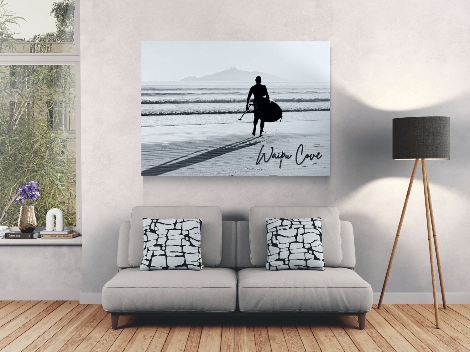 Black and white canvas print of a surfer on a beach at Waipa Cove, displayed on a living room wall above a gray sofa.